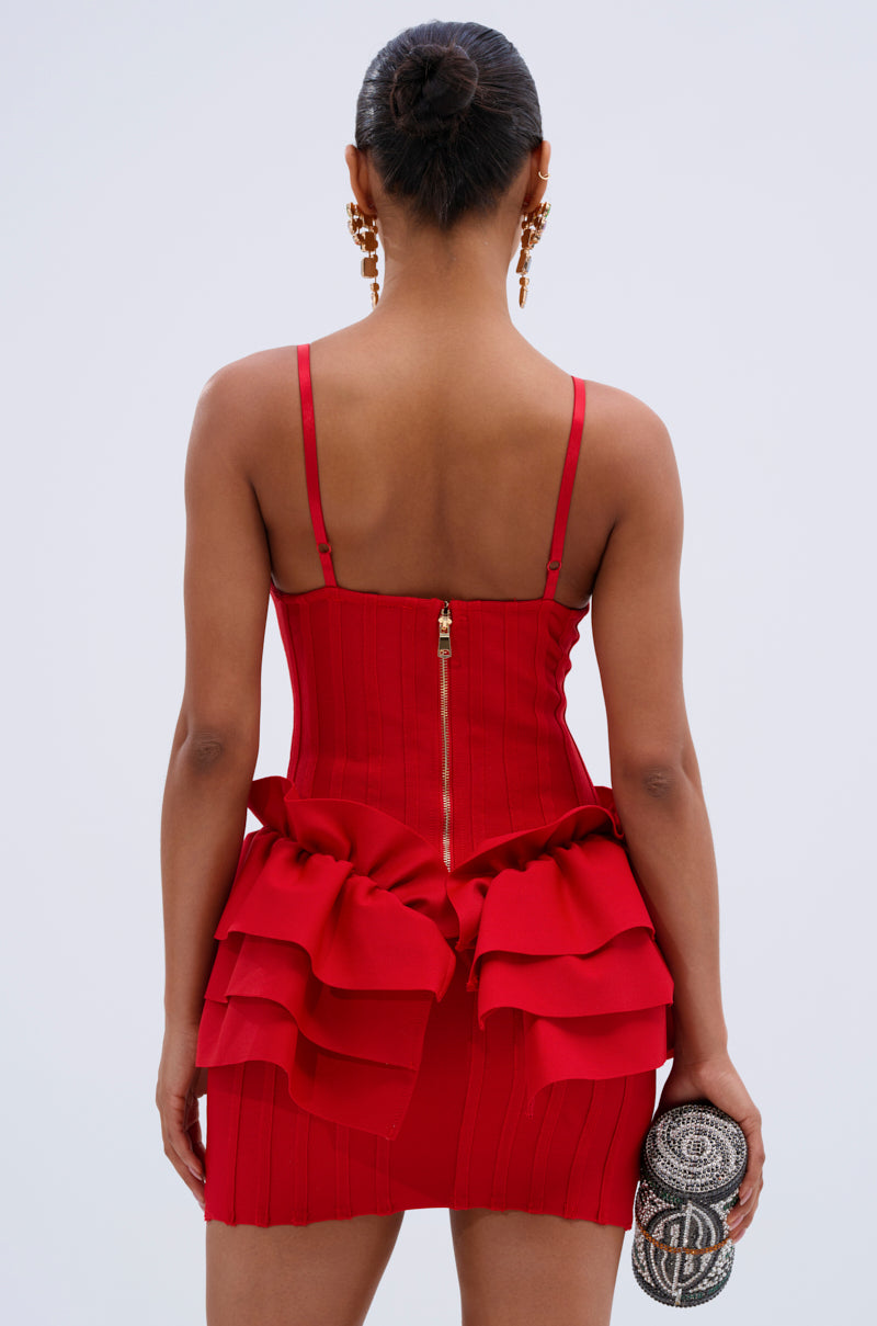 A woman facing away in the FELT CUTE RUFFLE BANDAGE MINI DRESS in red with a gold zipper, gold statement earrings, and a small round silver clutch. Her hair is styled in a neat bun.