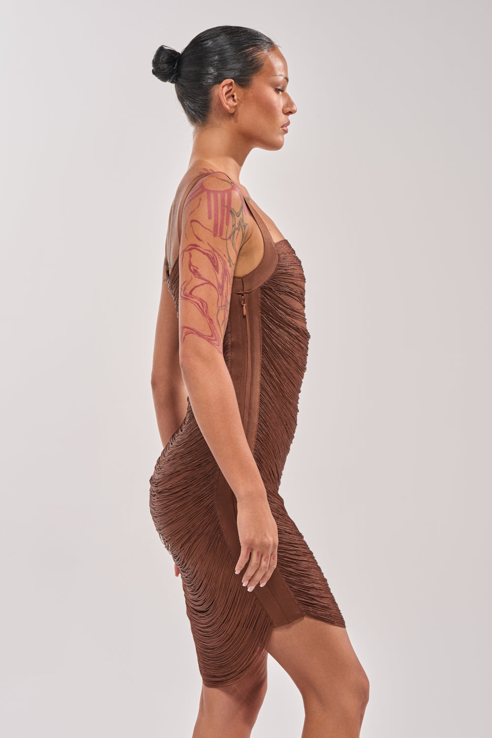 A person with dark hair in a bun and colorful arm tattoos is standing in profile, wearing the HYPNOTIZE ME FRINGE MINI DRESS IN BROWN against a plain light background. 3