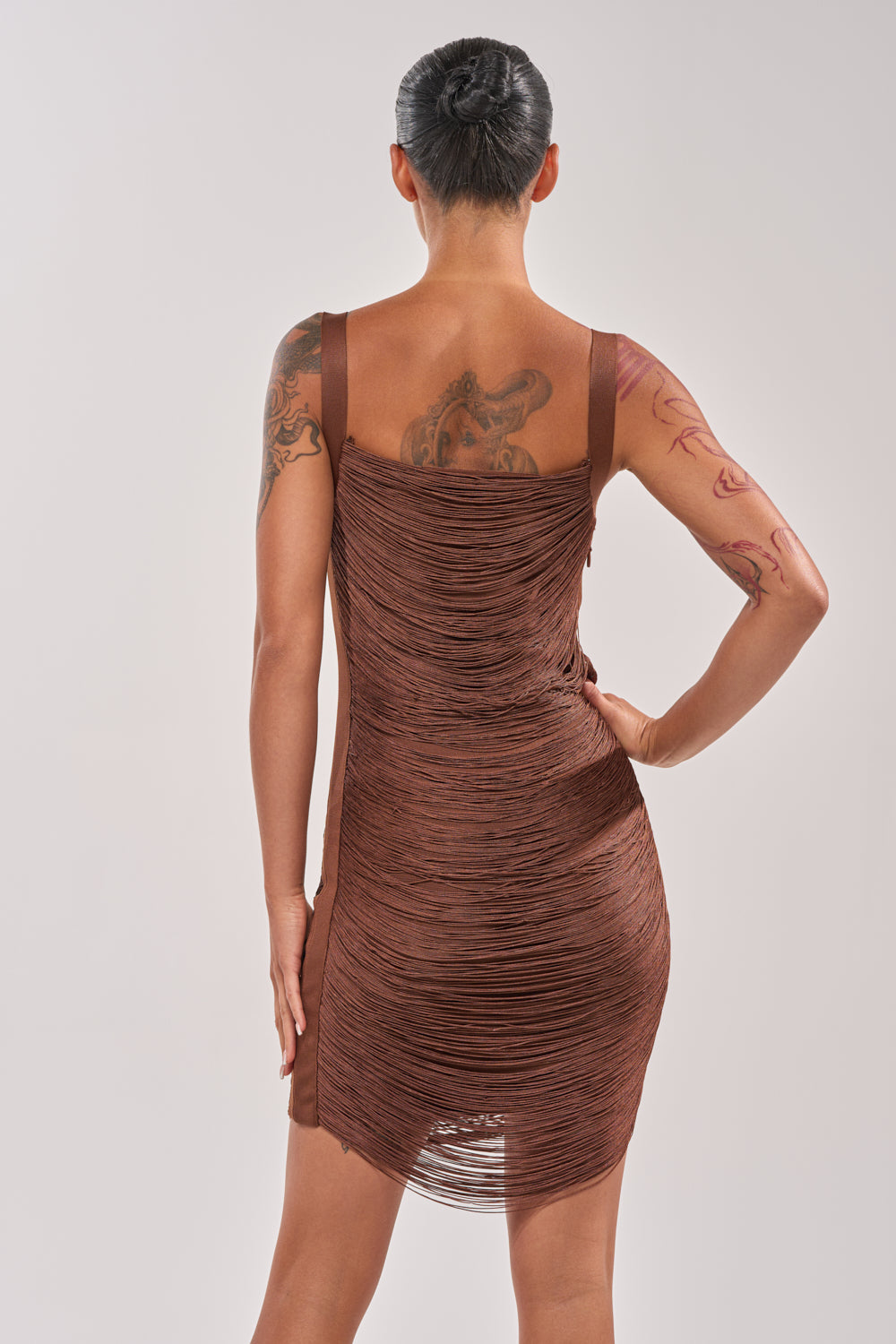 A model with dark hair in a bun showcases the HYPNOTIZE ME FRINGE MINI DRESS IN BROWN from the back, highlighting tattoos on their left arm and right shoulder, against a plain light background. 4