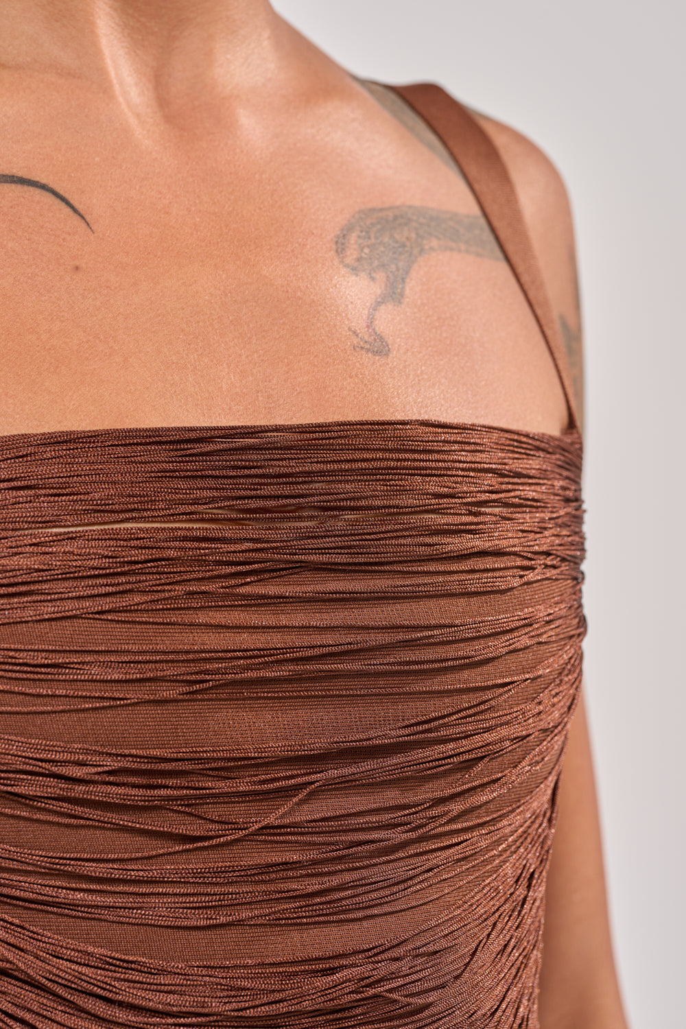 Close-up of a person wearing the HYPNOTIZE ME FRINGE MINI DRESS IN BROWN, featuring a textured, ruched design and thin straps. The upper chest and shoulder with a black tattoo near the collarbone are visible. 7