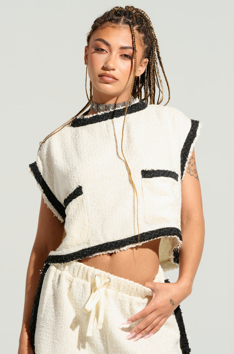 A person with long braided hair is wearing the EVERYONES TYPE SHORT SLEEVE TOP, a tweed-inspired off-white top with black trim and a front pocket. They are also sporting matching off-white pants with a drawstring. The person is posing against a plain, light-colored background. 0