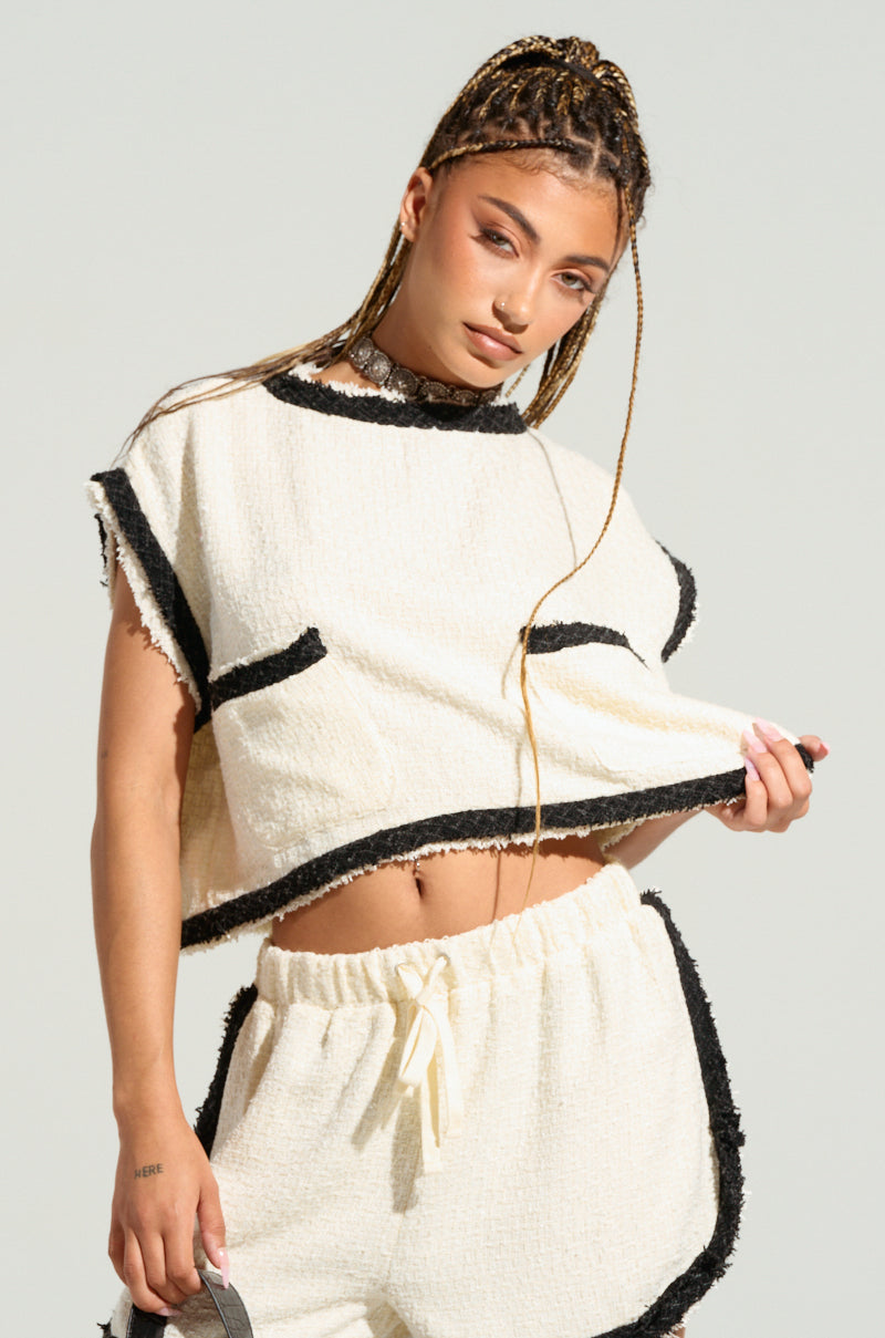 A person with long braided hair is posing in the EVERYONES TYPE SHORT SLEEVE TOP, a tweed-inspired two-piece cream-colored outfit with black trim. The top is slightly lifted, revealing their midriff, and they maintain a neutral facial expression. The background is plain and light-colored. 1