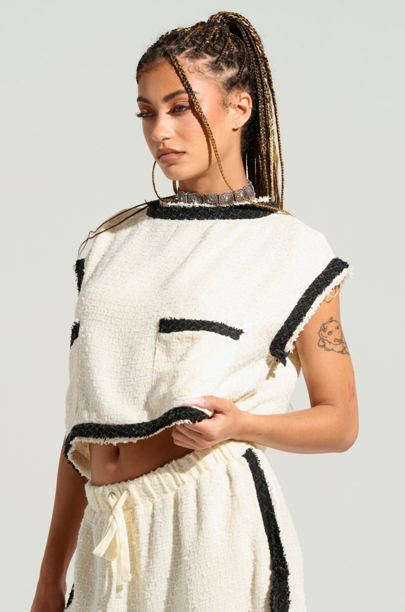A young woman with braided hair is wearing a chic, tweed-inspired sleeveless two-piece outfit featuring black trim, paired with the EVERYONES TYPE SHORT SLEEVE TOP. She has a tattoo on her upper arm and is looking down with a calm expression. The background is plain and light-colored, highlighting her fashion-forward choice. 2