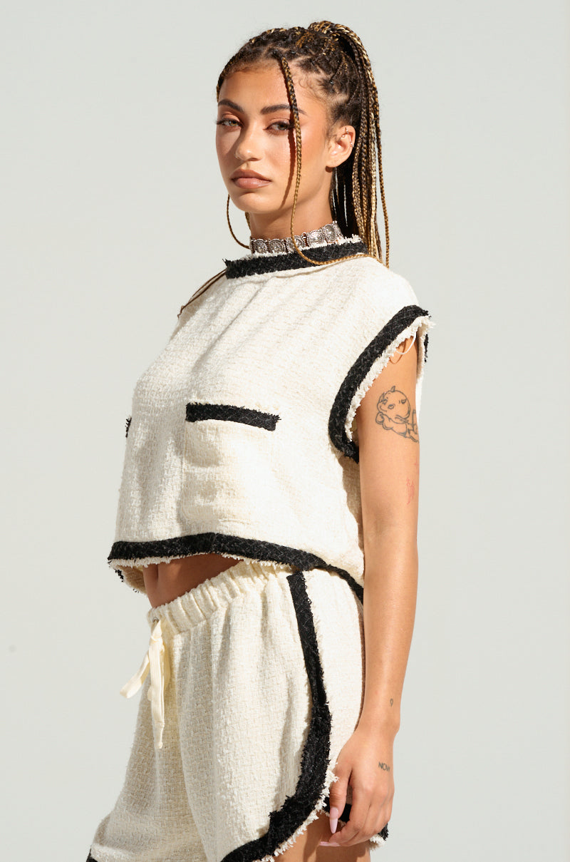A woman with long braids wears a two-piece set, including the EVERYONES TYPE SHORT SLEEVE TOP, which is a tweed-inspired sleeveless white top with black trim, paired with matching white shorts. She shows visible tattoos on her arm and maintains a neutral expression, all against a plain, light-colored background. 3