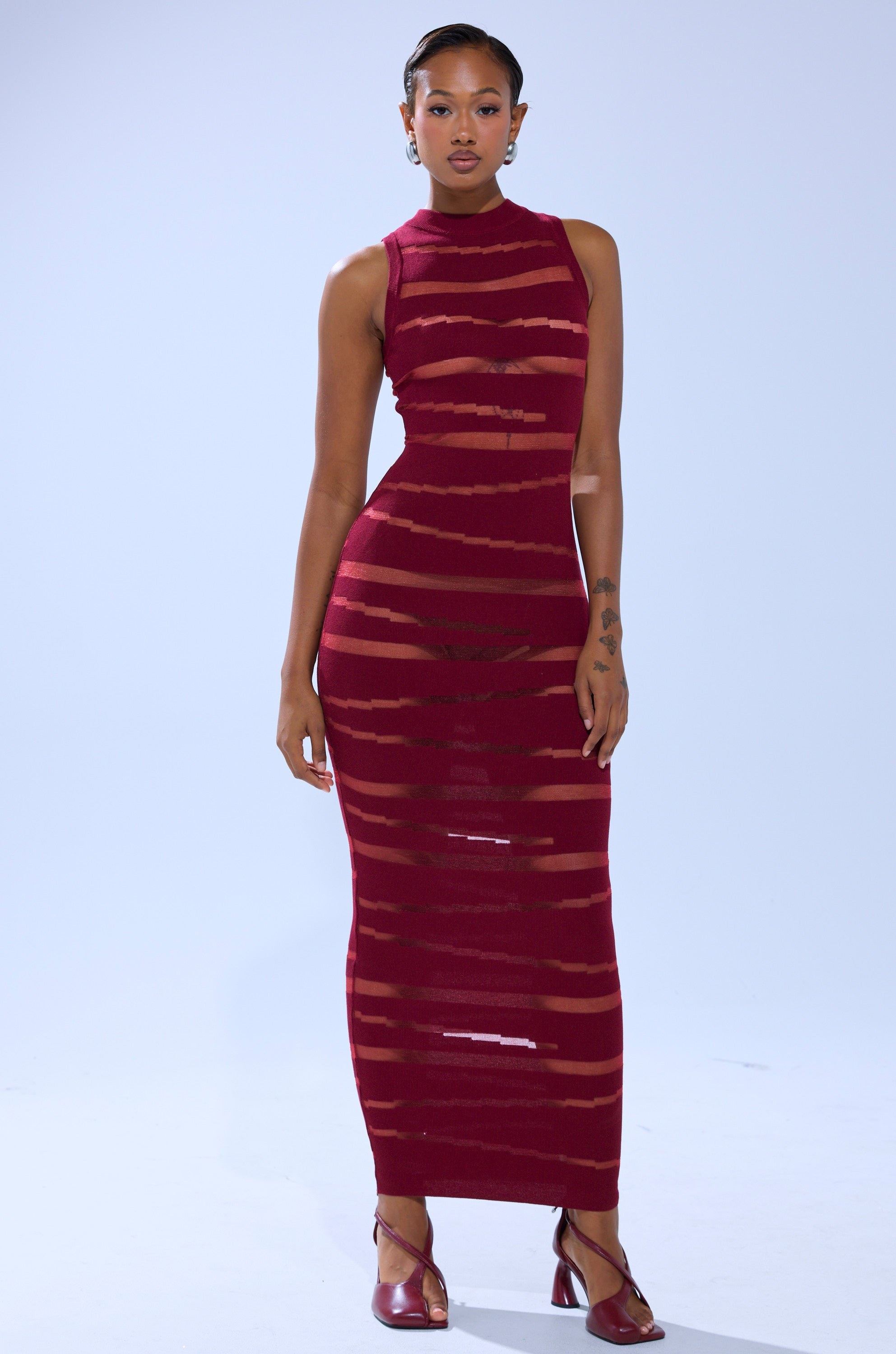 A woman stands against a plain light background wearing the CUT IT OUT SLEEVELESS KNIT MAXI DRESS IN BURGUNDY, a sleeveless knit maxi with horizontal sheer stripes, paired with matching heels.