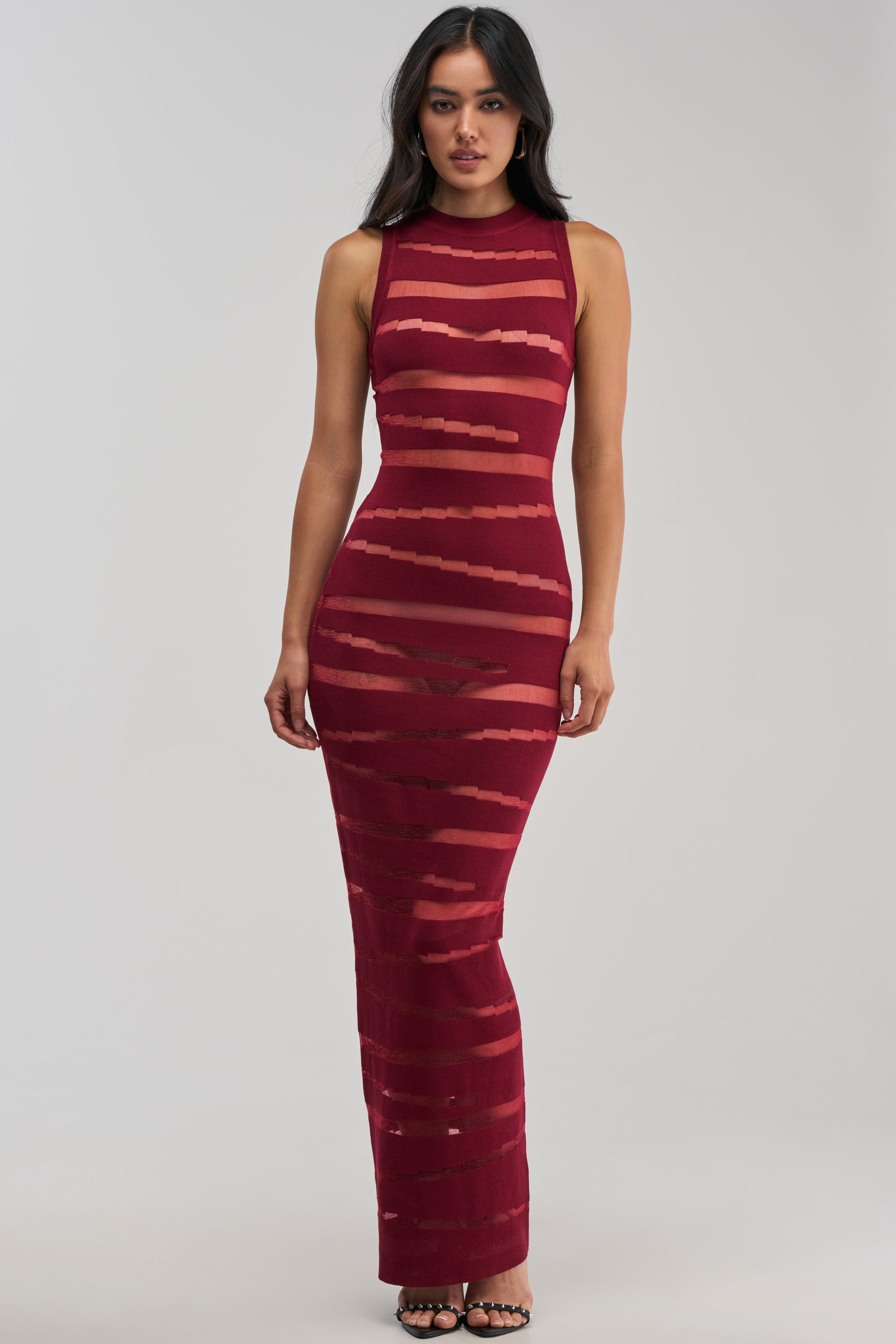 A woman wearing the CUT IT OUT Sleeveless Knit Maxi Dress in Burgundy—a fitted, floor-length cocktail dress featuring horizontal sheer stripes—stands against a plain light background.