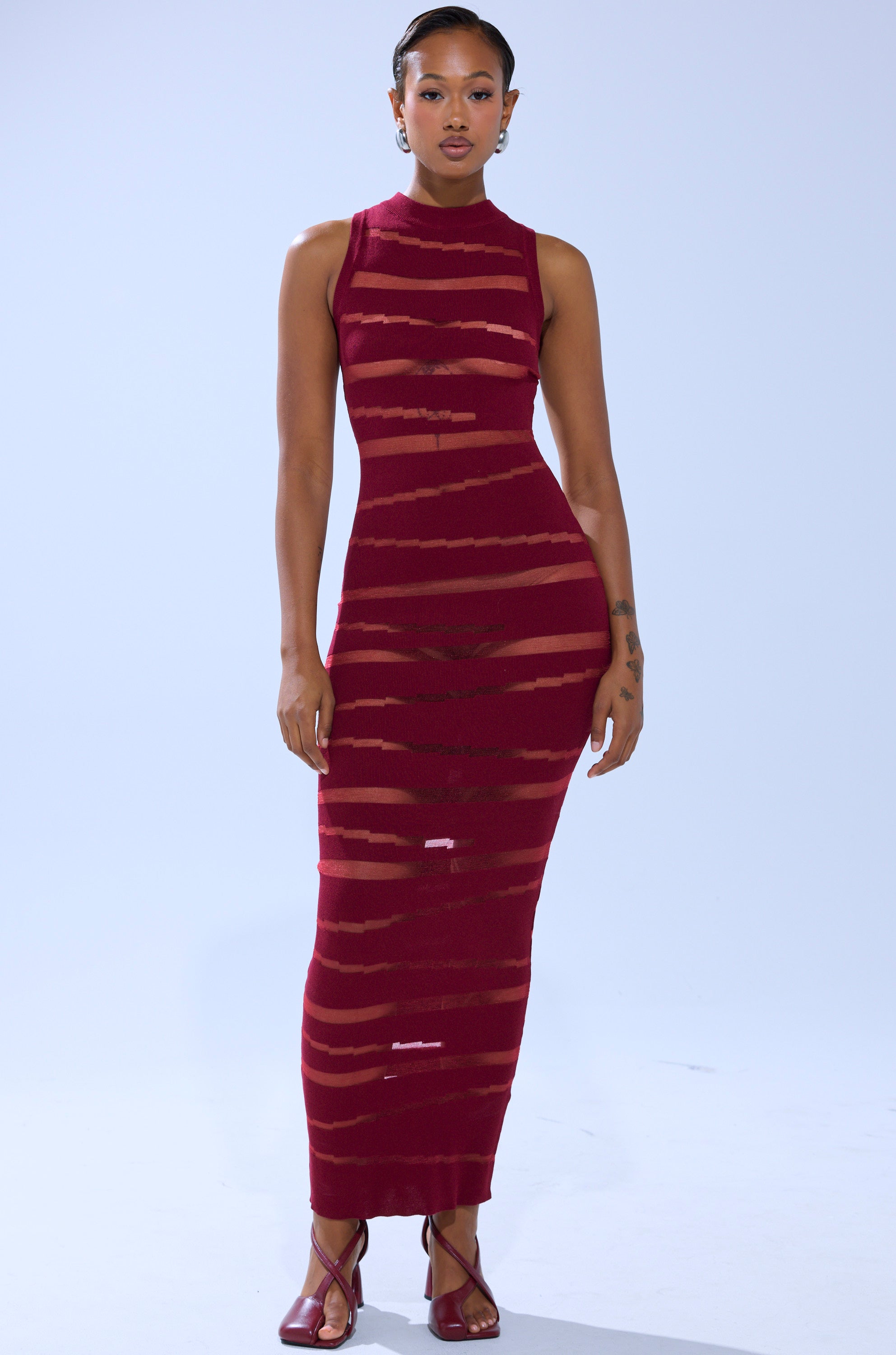 A woman with short hair and minimal jewelry poses against a plain background in the CUT IT OUT SLEEVELESS KNIT MAXI DRESS IN BURGUNDY, featuring sheer horizontal stripes, cut-out paneling, and paired with open-toe heels.
