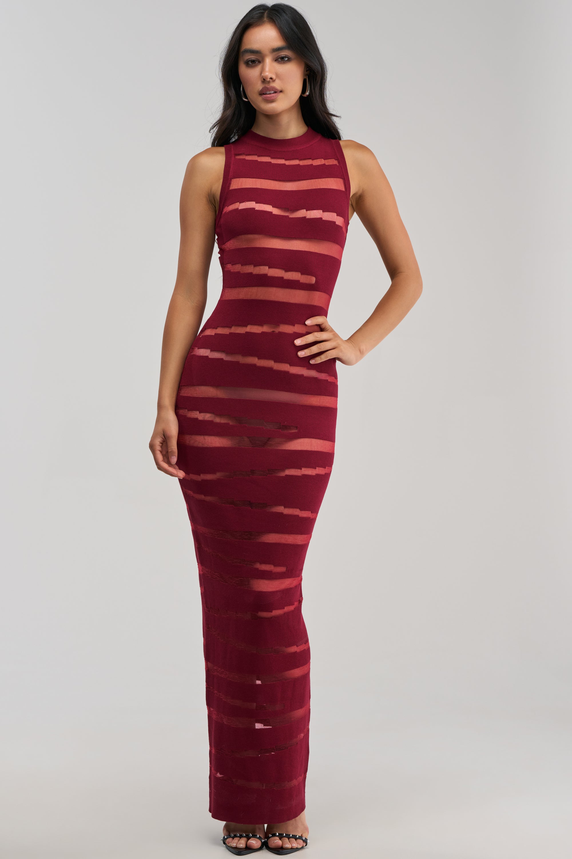 A woman poses with one hand on her hip against a plain light background, wearing the CUT IT OUT Sleeveless Knit Maxi Dress in Burgundy—a floor-length dress featuring horizontal sheer cutouts.