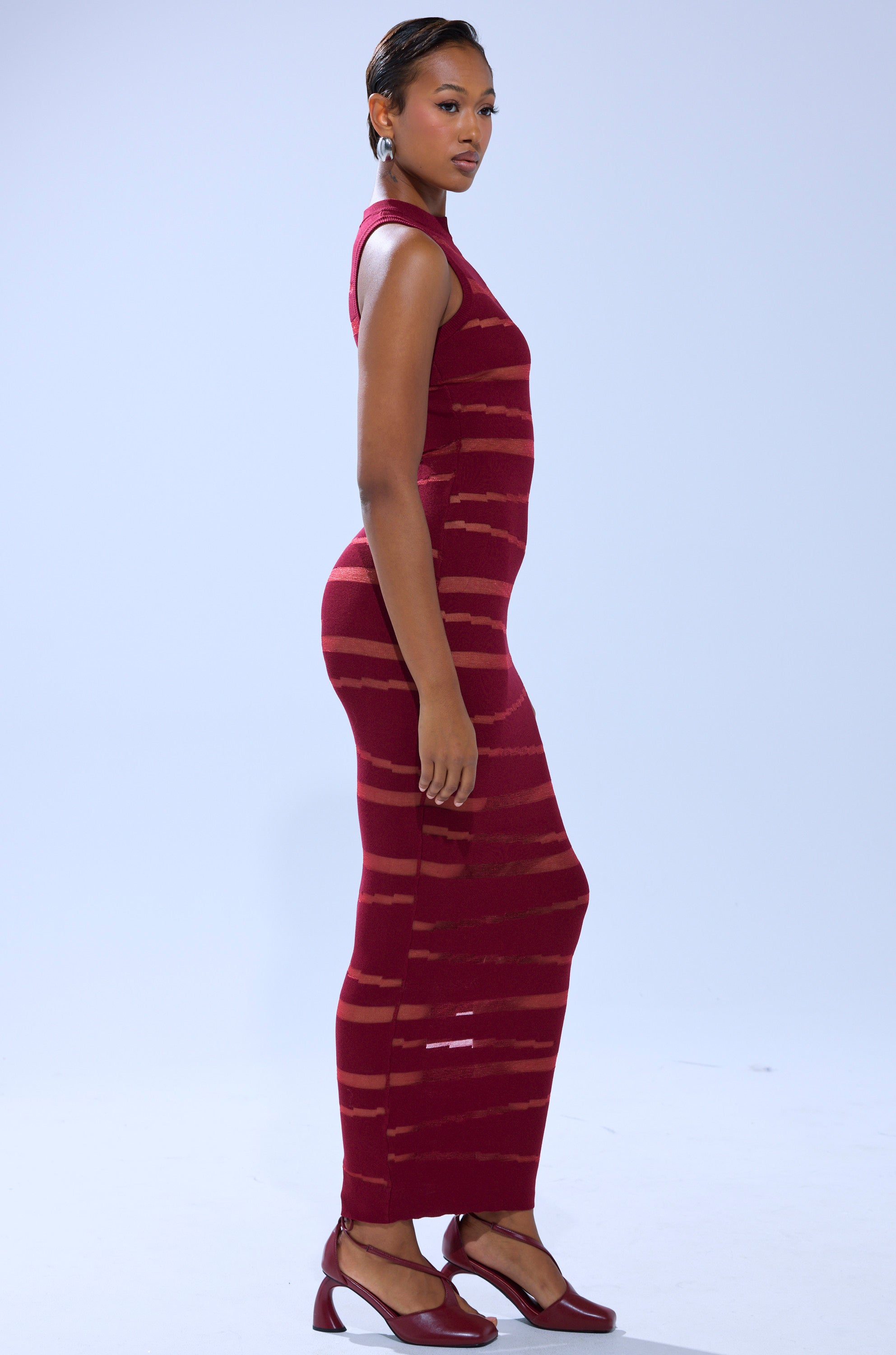 A woman stands sideways in the CUT IT OUT SLEEVELESS KNIT MAXI DRESS IN BURGUNDY, an ankle-length knit dress with horizontal sheer stripes and cut-out paneling. She pairs it with red heels against a plain light background.