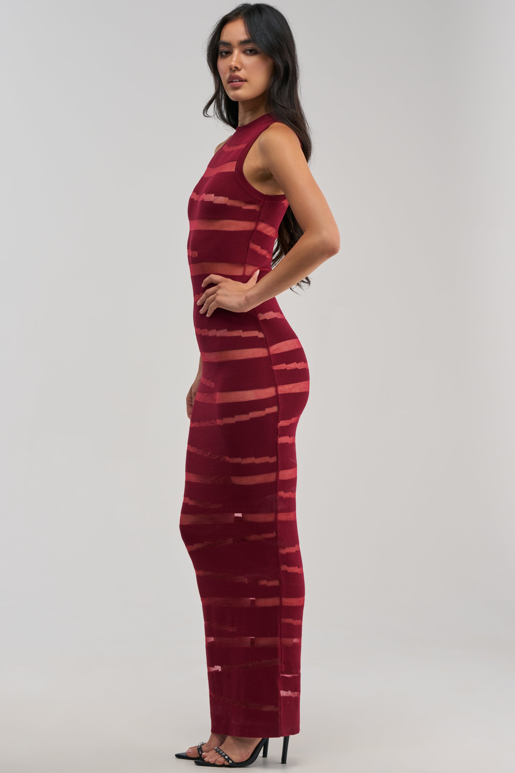 A woman stands sideways in black high-heeled sandals against a plain light background, wearing the CUT IT OUT SLEEVELESS KNIT MAXI DRESS IN BURGUNDY—a sleeveless, form-fitting maxi dress featuring horizontal sheer stripes. This sexy cocktail party dress is perfect for any evening out.