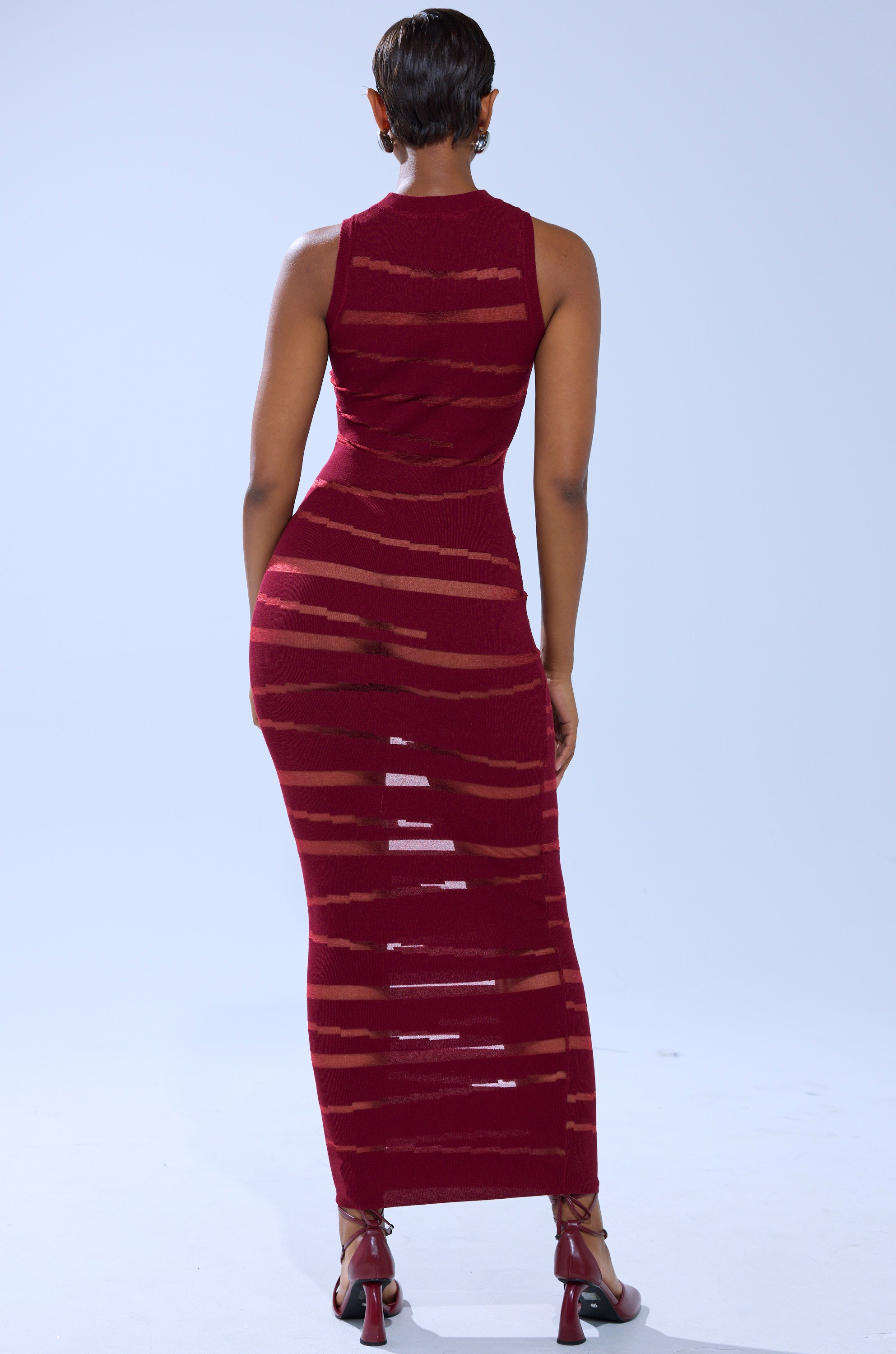 A person with short hair wears the CUT IT OUT SLEEVELESS KNIT MAXI DRESS IN BURGUNDY, an ankle-length, form-fitting dress with sheer cut out panels, styled with red high-heeled sandals and standing facing away.
