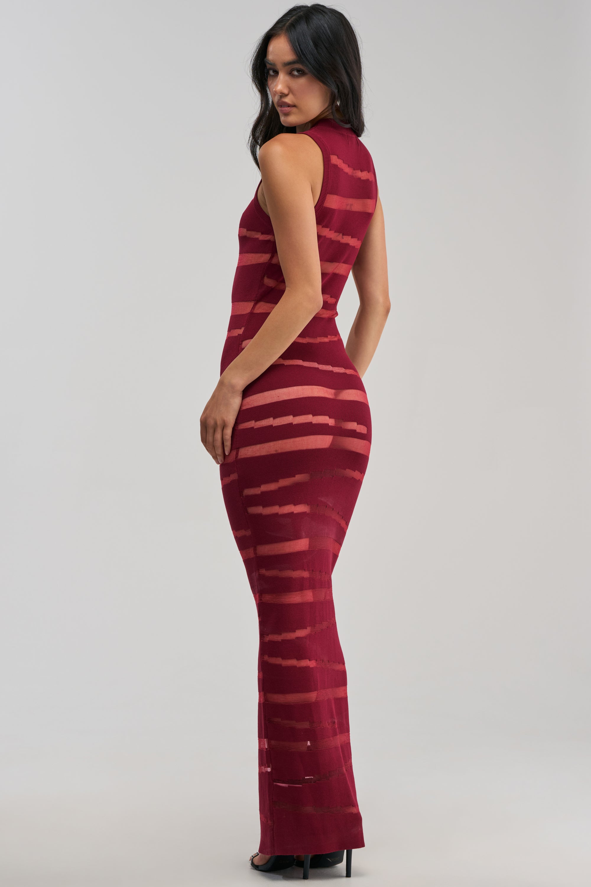 A woman wearing the “CUT IT OUT Sleeveless Knit Maxi Dress in Burgundy”—a sleek, body-hugging maroon dress with horizontal stripe detailing—stands in high heels, gazing over her shoulder against a simple light backdrop.
