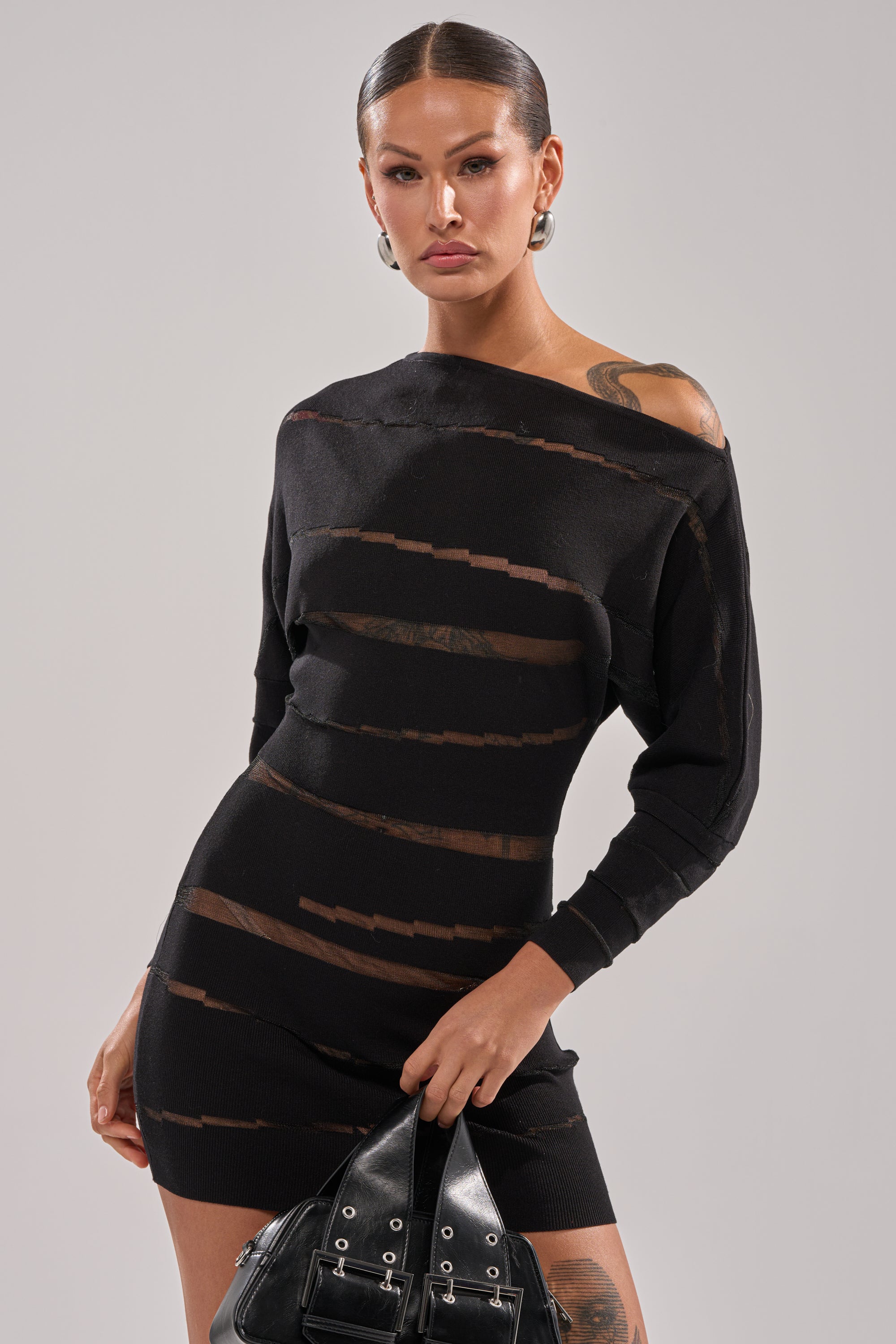 Model exudes confidence in the AFTER DARK STRIPED MINI DRESS, featuring a sheer mesh, one-shoulder design. She pairs it with a black handbag with silver buckles, slicked-back hair, and large hoop earrings for a bold look.