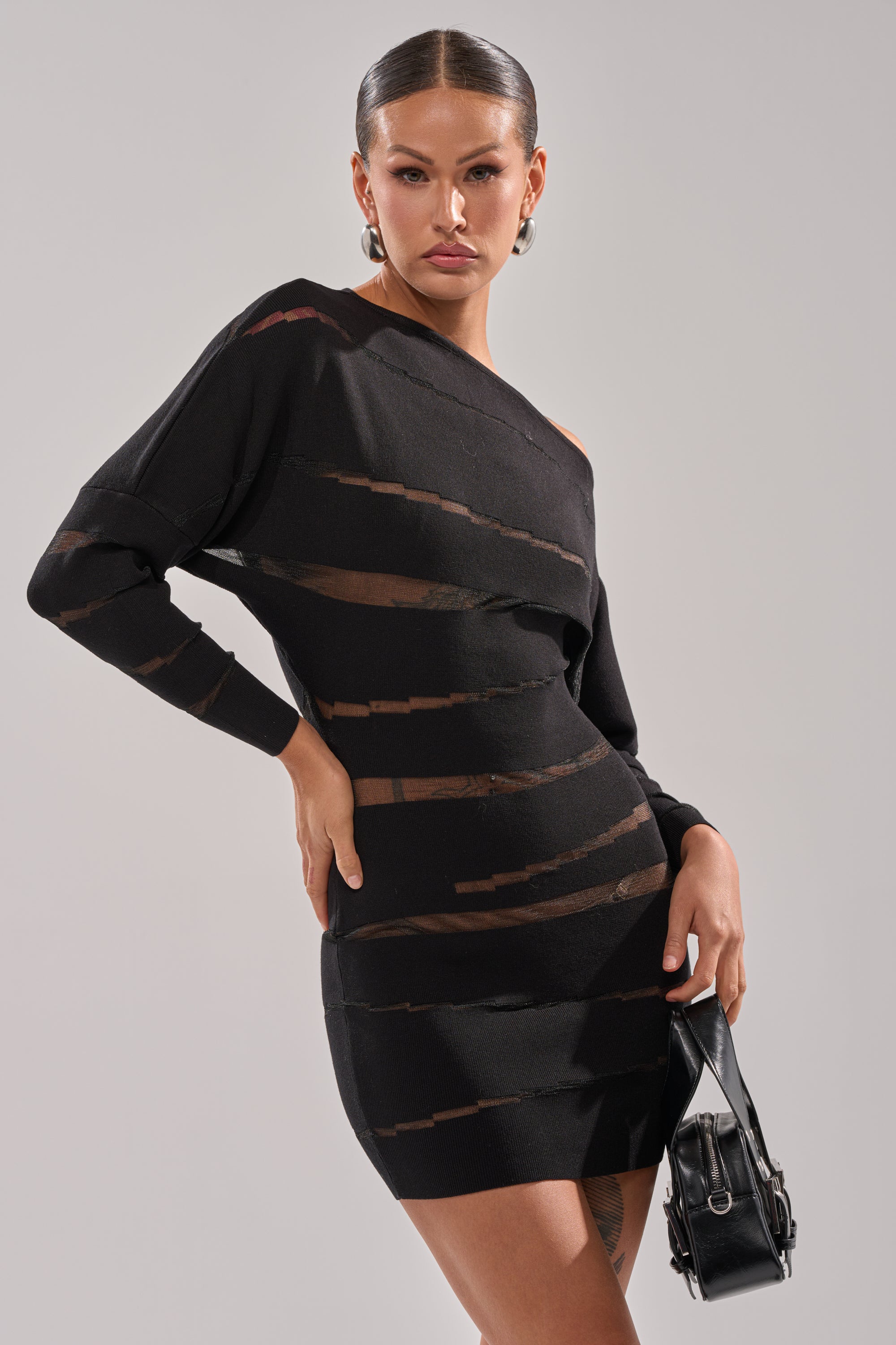 A woman poses confidently in the AFTER DARK STRIPED MINI DRESS, featuring sheer mesh panels, with slicked-back hair, hoop earrings, and a black handbag against a plain gray background.