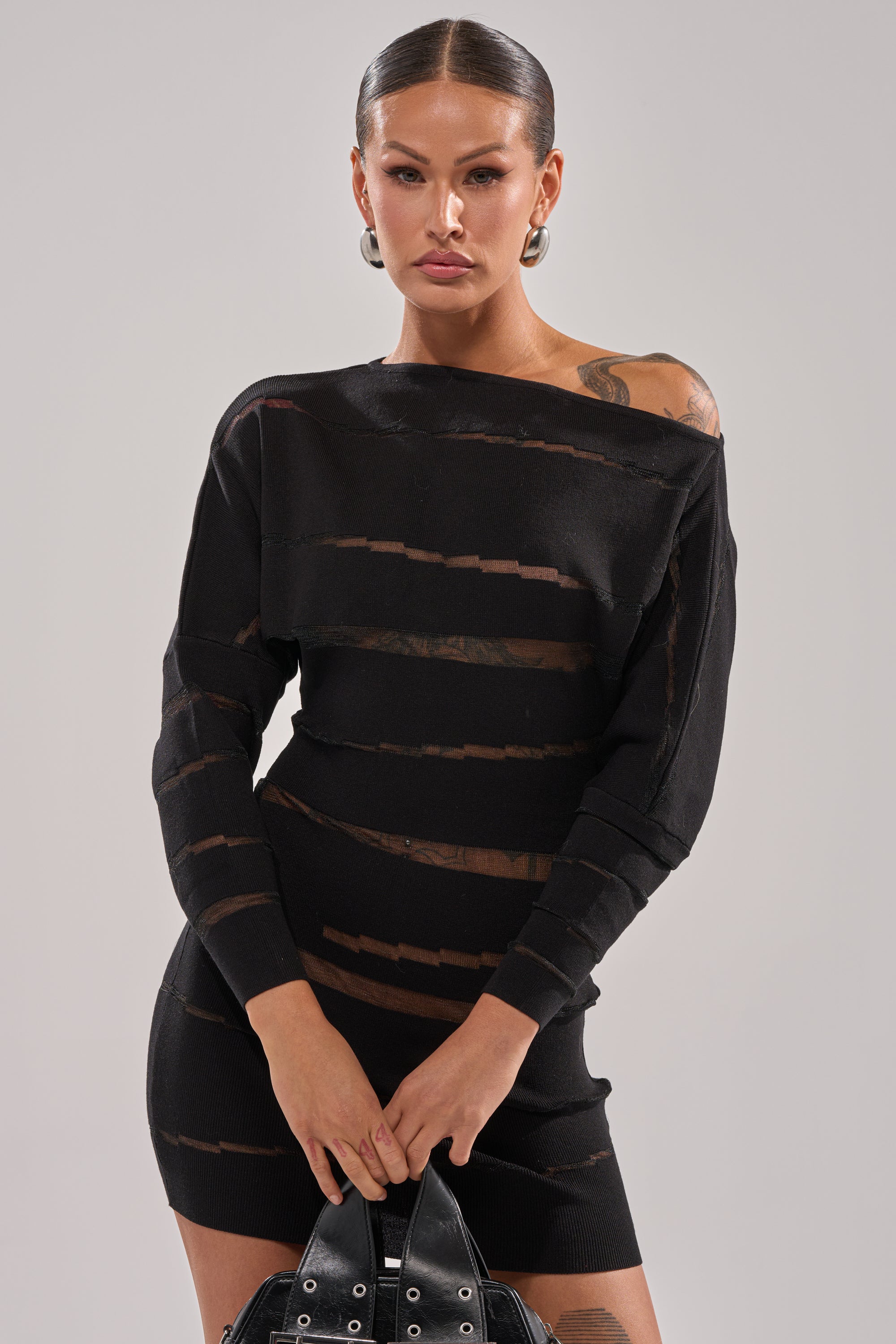 A woman with slicked-back hair models the AFTER DARK STRIPED MINI DRESS, a black sheer mesh striped mini dress. She accessorizes with hoop earrings, a black bag, and displays tattoos, posing against a plain light grey background.