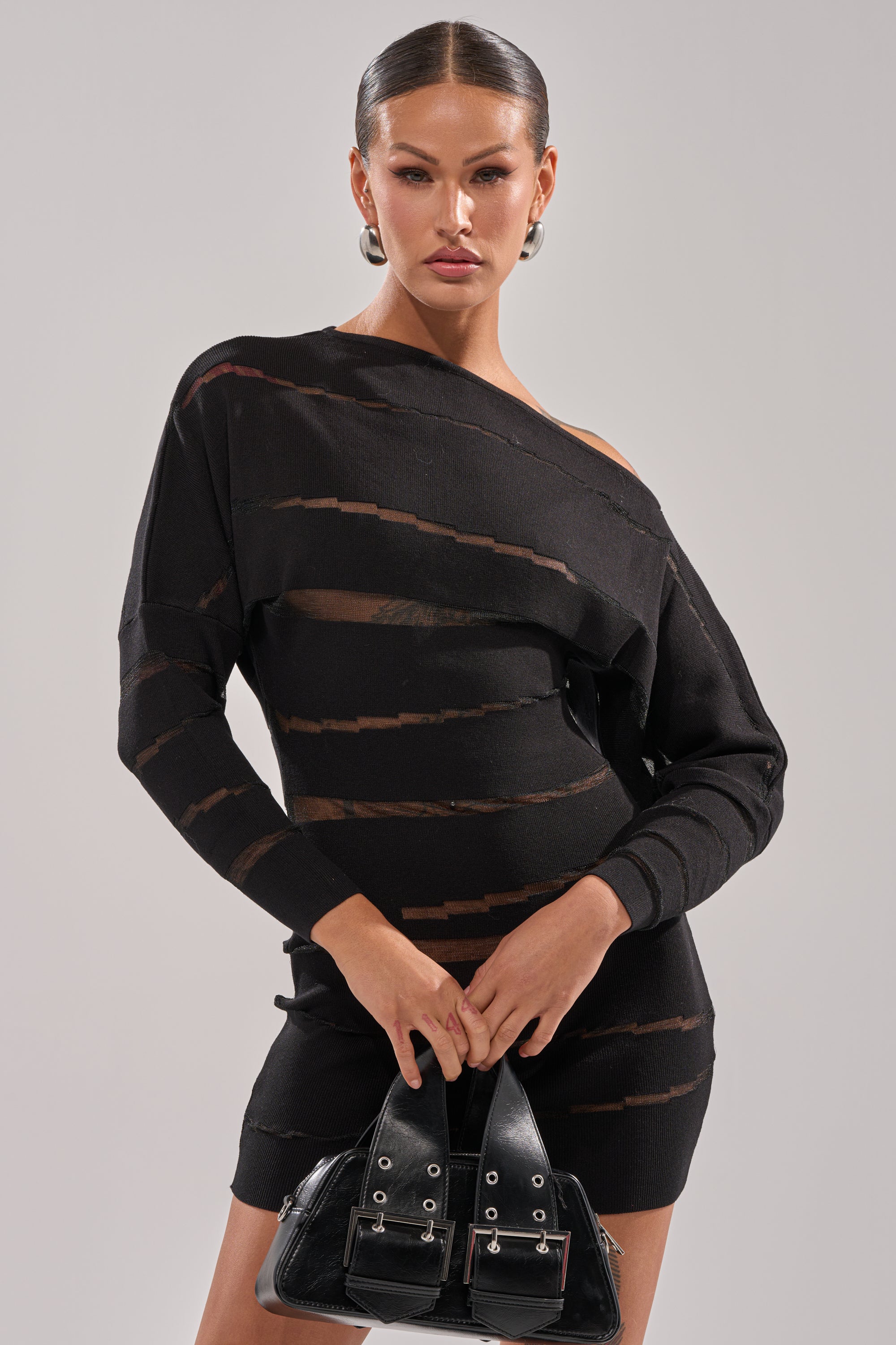 A woman with slicked-back hair wears the AFTER DARK STRIPED MINI DRESS—an asymmetrical black sheer mesh mini with horizontal stripes—and holds a black handbag with silver buckles against a plain light background.