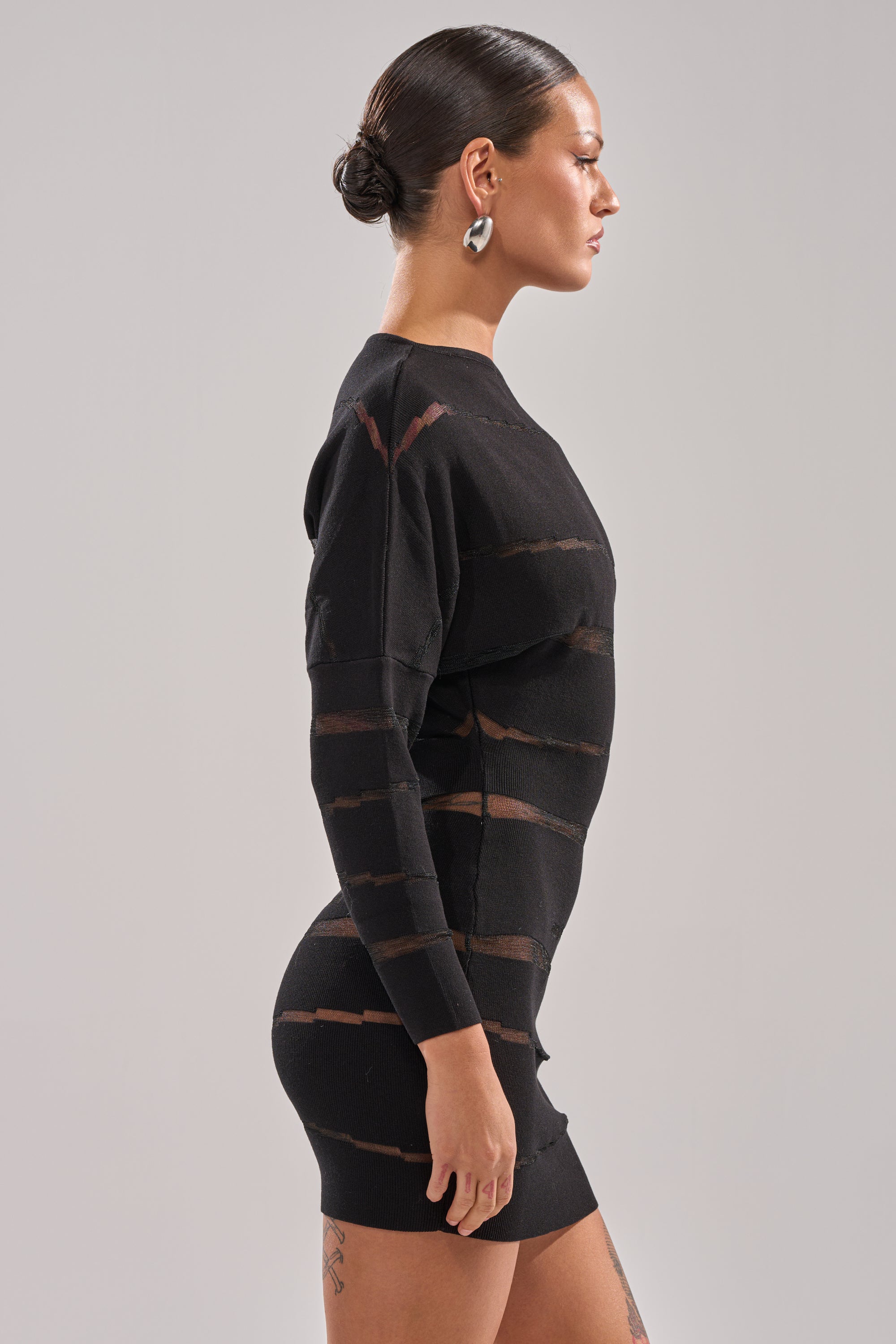 A woman with slicked-back hair and silver hoop earrings stands in profile against a light gray background, wearing the AFTER DARK STRIPED MINI DRESS—a fitted black dress featuring sheer horizontal stripes.