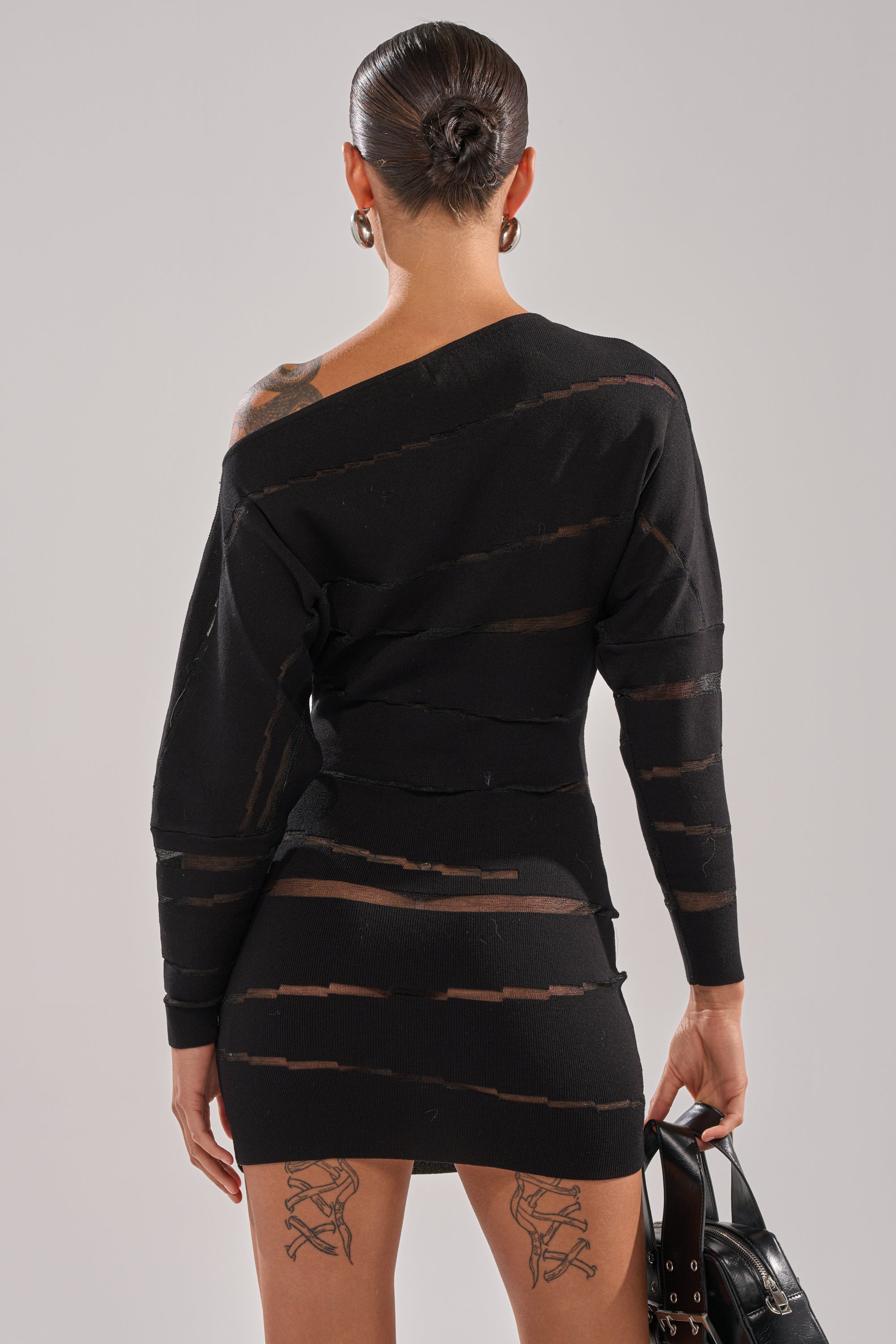 Wearing the AFTER DARK STRIPED MINI DRESS—an asymmetrical, curve-hugging black mini with sheer mesh stripes—someone with slicked-back hair and visible thigh tattoos holds a black handbag, facing away from the camera.