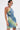 Wearing the FUN N FLIRTY PRINTED MINI DRESS with a blue-green pattern, they carry a silver handbag, sport hoop earrings, show a tattoo on their upper arm, have short dark hair, and display a neutral expression.
