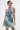 A person with short dark hair and a blonde streak models the FUN N FLIRTY PRINTED MINI DRESS, featuring a blue-green rippled print halter style, visible tattoos, and a shiny small handbag against a plain white background.