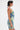 A person with short dark hair wears the FUN N FLIRTY PRINTED MINI DRESS—a sleeveless blue-green print mini dress with ruching—paired with a shiny silver clutch, red abstract shoulder tattoo visible, against a plain white background.