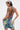 A person with short, two-toned hair and visible tattoos models the FUN N FLIRTY PRINTED MINI DRESS—a backless blue-green halter mini dress with yellow pleats—while holding a silver clutch against a white background.