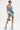 A person models the FUN N FLIRTY PRINTED MINI DRESS—a blue-green ruched halter mini dress—paired with a small silver purse and light blue fluffy shoes, showcasing arm and thigh tattoos against a white background.