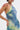 A woman poses confidently with one hand on her hip, wearing the FUN N FLIRTY PRINTED MINI DRESS—a ruffled, blue-green print mini dress with a halter neckline. She has short hair, statement earrings, and a pink abstract design on her arm against a white background.