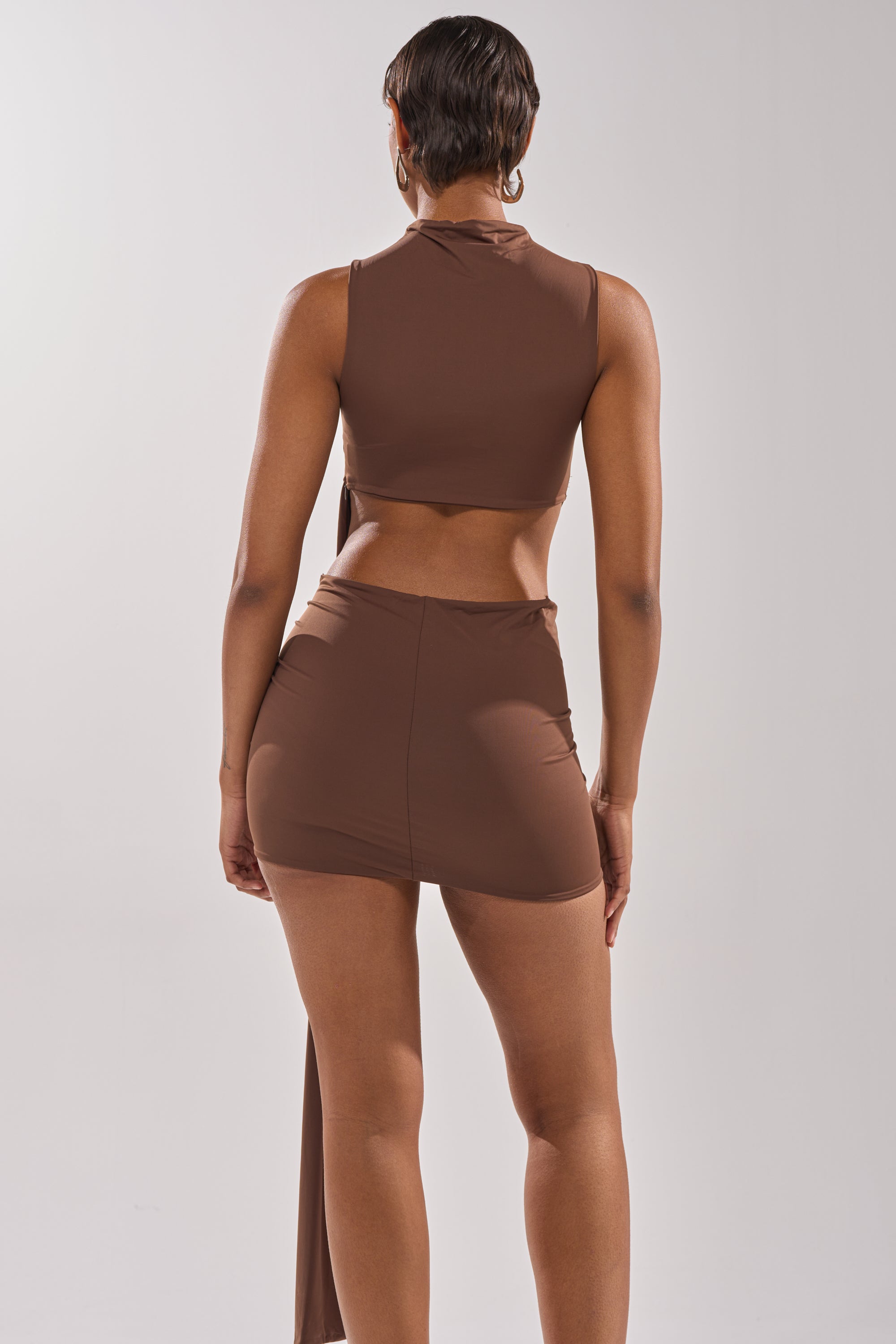 A woman stands facing away from the camera, wearing the HOTTEST IN THE ROOM MINI DRESS in brown. The sleeveless, body-hugging dress features a high-waisted fit and a mini length. The background is plain and light-colored.