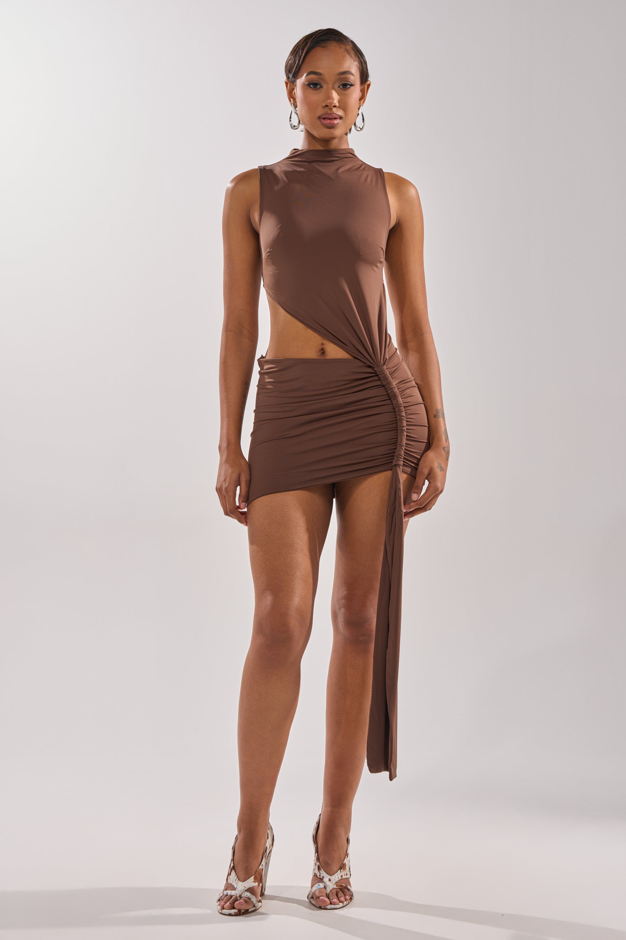A woman with short hair, hoop earrings, and open-toe heels poses against a plain background wearing the HOTTEST IN THE ROOM MINI DRESS—a brown, body-hugging mini dress featuring asymmetrical ruched detailing and cut-out accents.