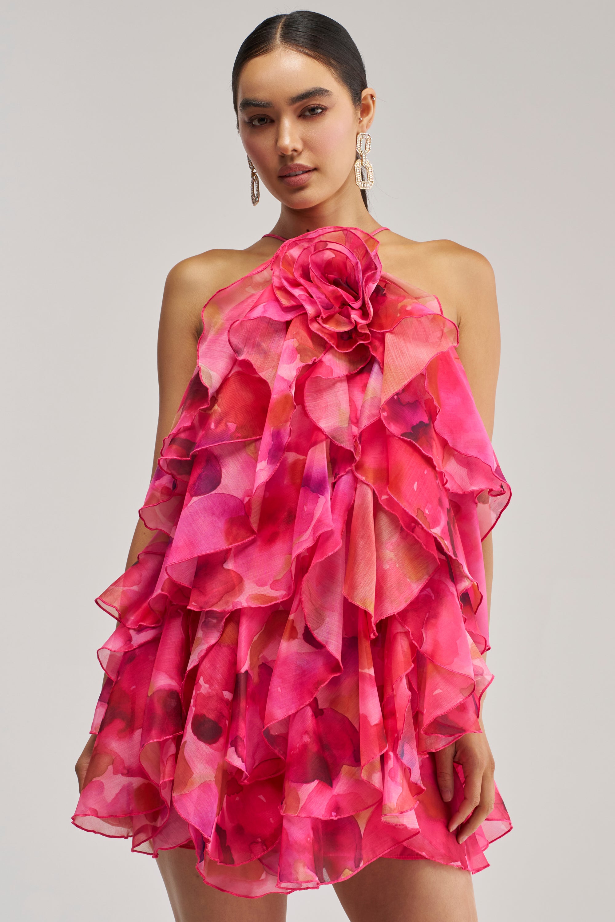 A woman stands facing forward in the I AM THE PARTY MINI DRESS IN POP PINK, featuring bright pink ruffle tiers and a bold floral detail at the neckline. With her dark hair pulled back and dangling earrings, she perfectly showcases this statement-making special occasion dress.