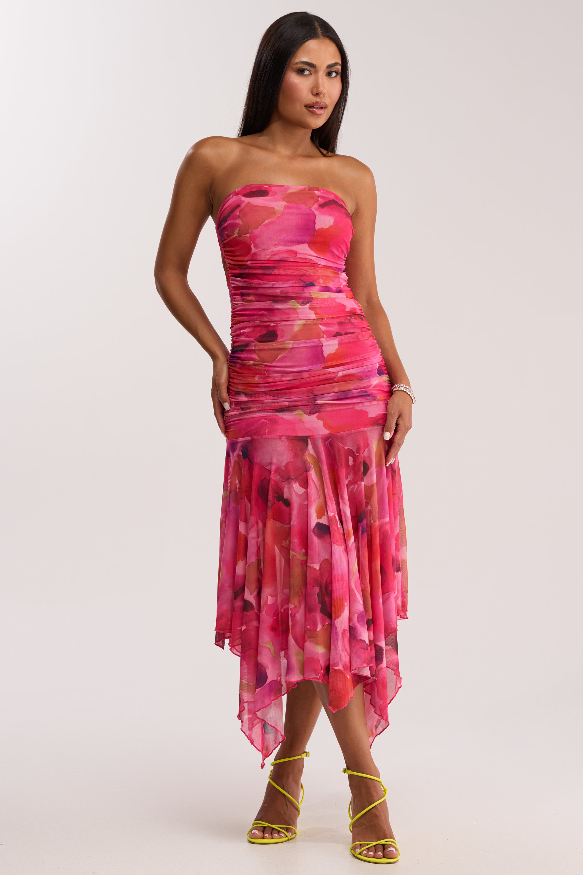 Wearing the BRUNCH DATE MESH MIDI DRESS IN PINK MULTI with a ruched bodice and asymmetrical hem, a woman stands confidently in yellow strappy heels, her straight dark hair contrasting with the dress's abstract print and the light background.