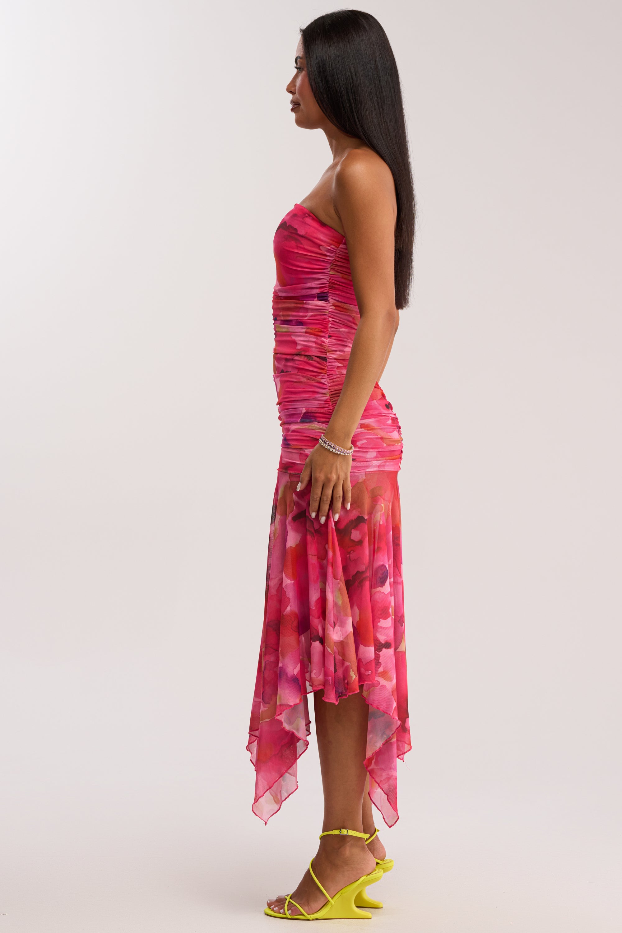 A woman in profile wears the BRUNCH DATE MESH MIDI DRESS IN PINK MULTI, featuring a strapless ruched mesh design with a floral asymmetrical hem. She pairs it with yellow strappy heels and a bracelet against a plain light background.