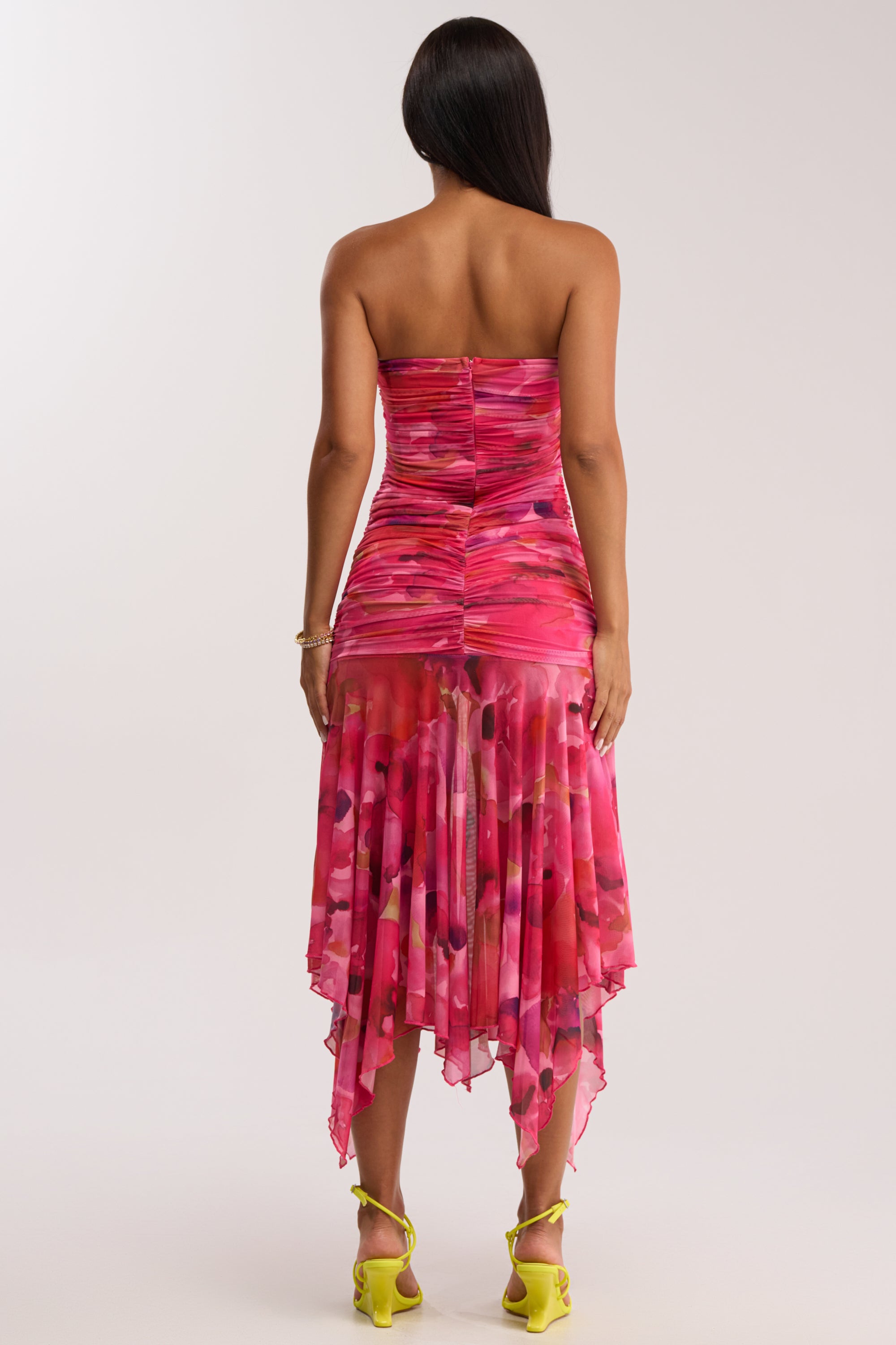 A woman with long dark hair faces away, wearing the BRUNCH DATE MESH MIDI DRESS IN PINK MULTI—an abstract floral, pink strapless midi dress with ruched mesh and a handkerchief hemline—paired with yellow heels.