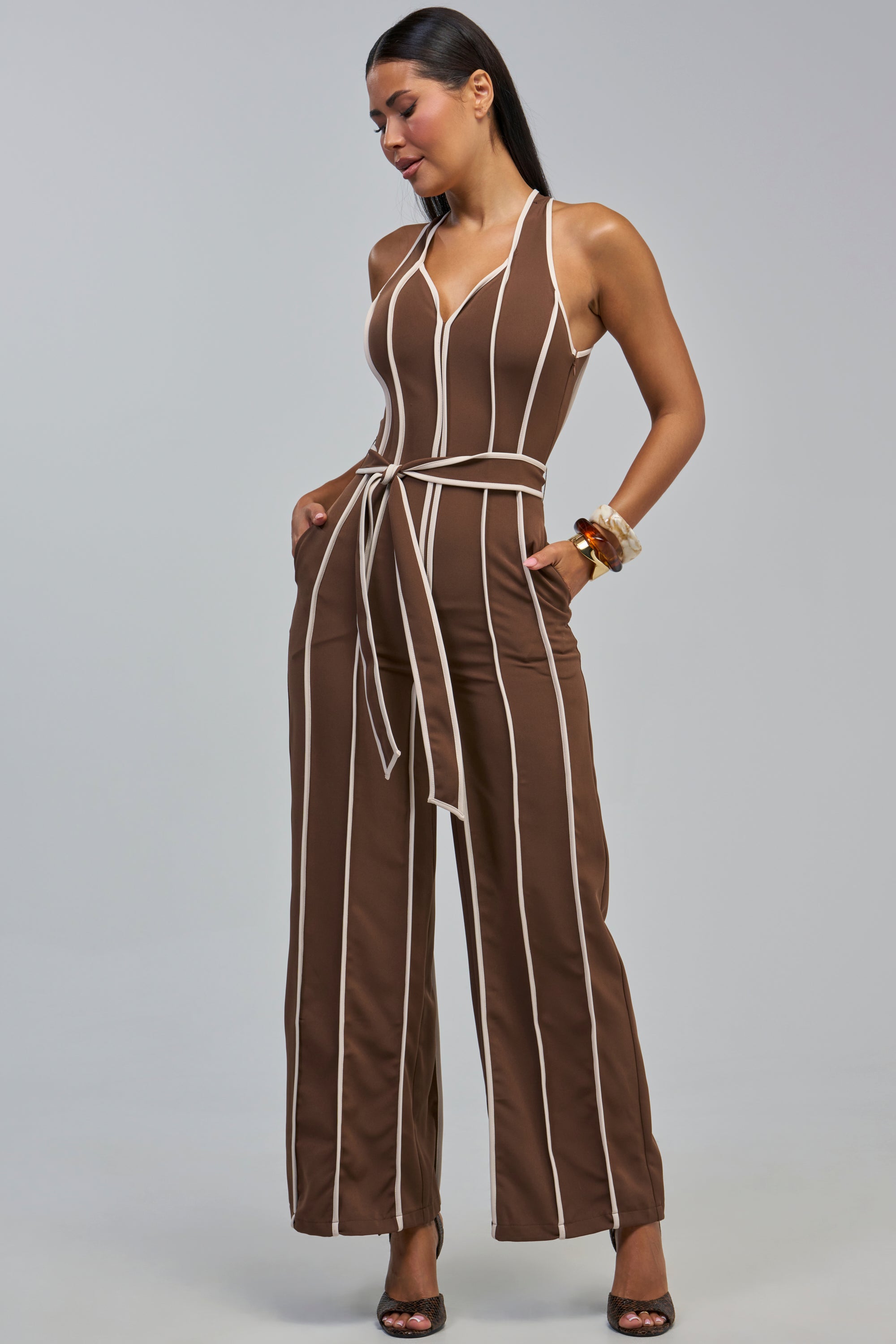A woman stands with her hands in the pockets of the SUMMER SERENADE STRIPE HALTER JUMPSUIT IN BROWN, which features white vertical stripes and a matching belt, looking down against a plain gray background.