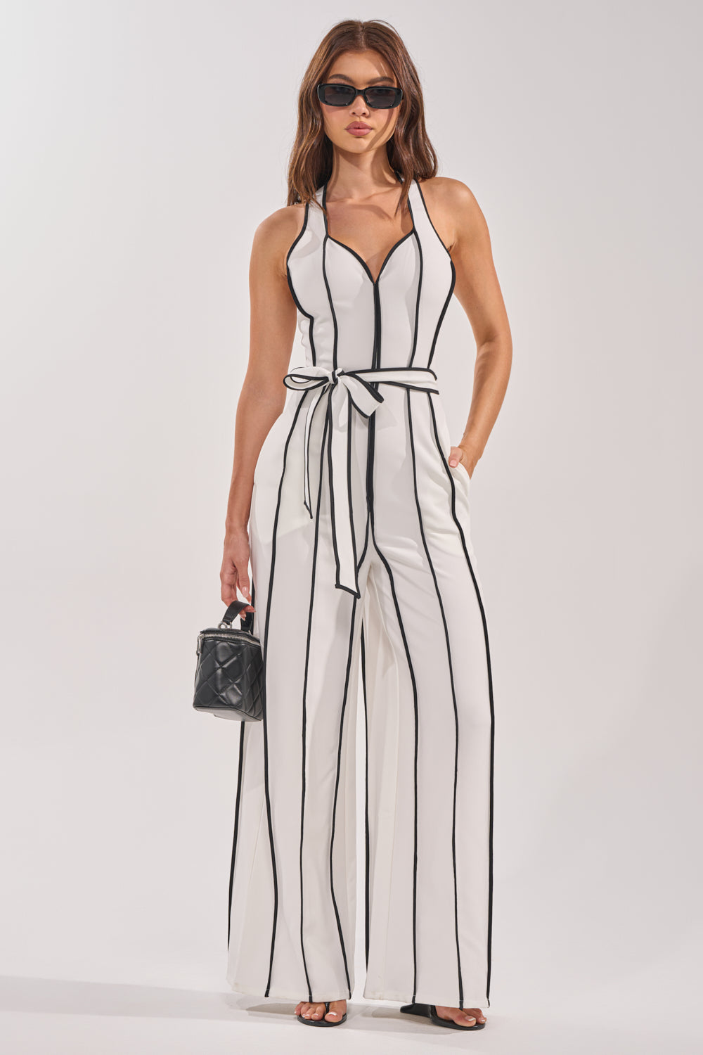 A woman models the SUMMER SERENADE STRIPE HALTER JUMPSUIT—a sleeveless white striped piece with a belted waist and wide legs. She pairs it with sunglasses, carries a black handbag, and poses against a plain light background. 0