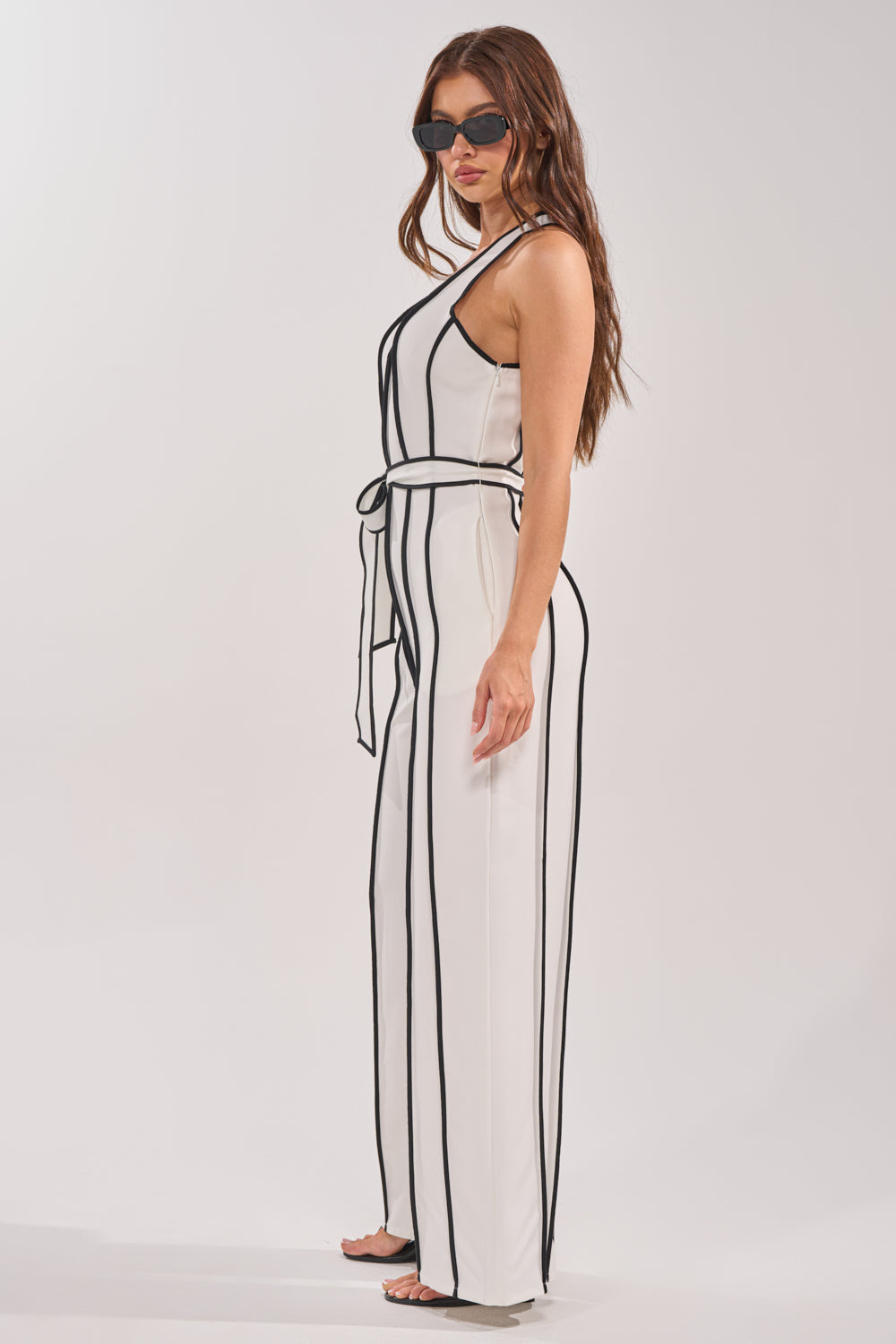 Woman wearing sunglasses and the SUMMER SERENADE STRIPE HALTER JUMPSUIT—a sleeveless white jumpsuit with vertical black stripes and matching belt—stands sideways against a plain light background. 2
