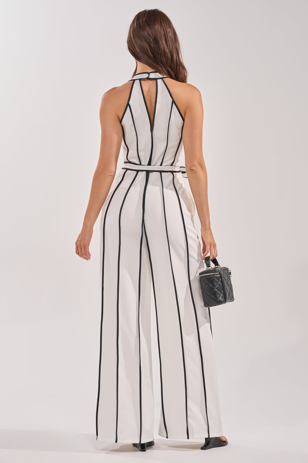A woman faces away, wearing the SUMMER SERENADE STRIPE HALTER JUMPSUIT with white and black vertical stripes and wide legs. She holds a small black quilted handbag, her long brown hair cascading down against a light background. 3