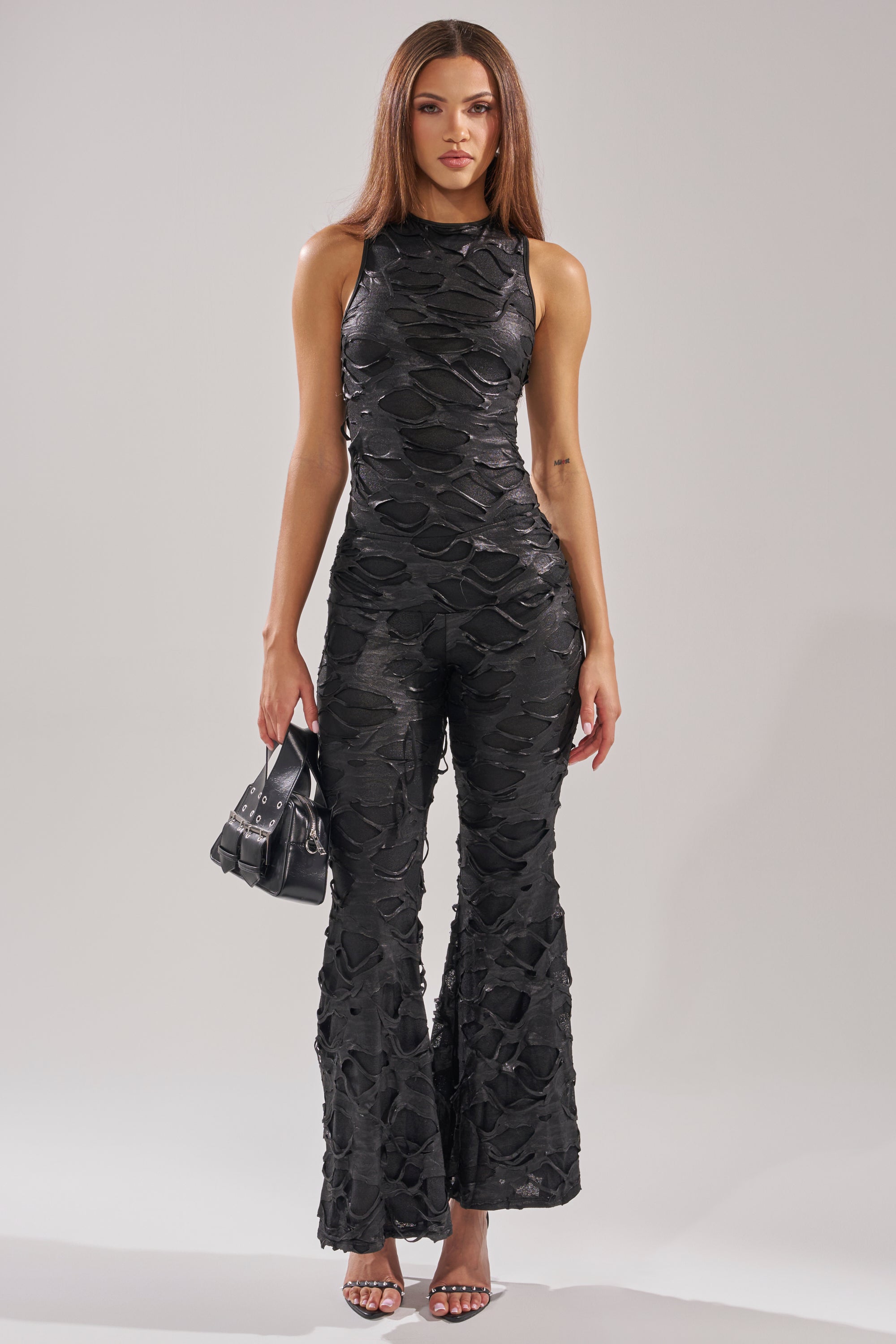 A woman with straight brown hair and a neutral expression stands against a plain background, wearing the SASHA LACE SLEEVELESS JUMPSUIT in black with flared legs, silver high-heeled sandals, and holding a small black handbag.
