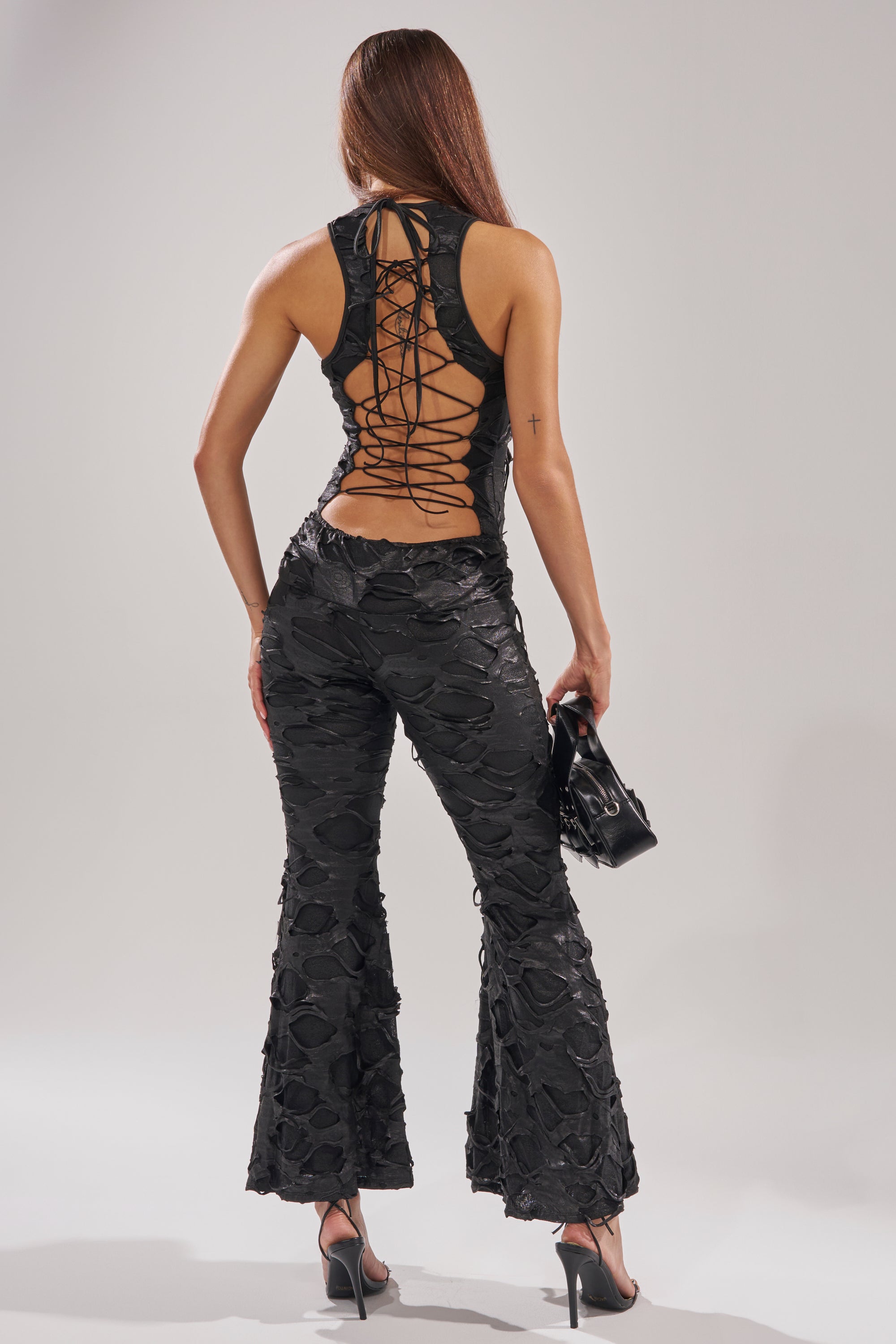 A woman faces away, wearing the SASHA LACE SLEEVELESS JUMPSUIT with a lace-up back and flared legs. She carries a small black handbag and pairs the outfit with high-heeled sandals.