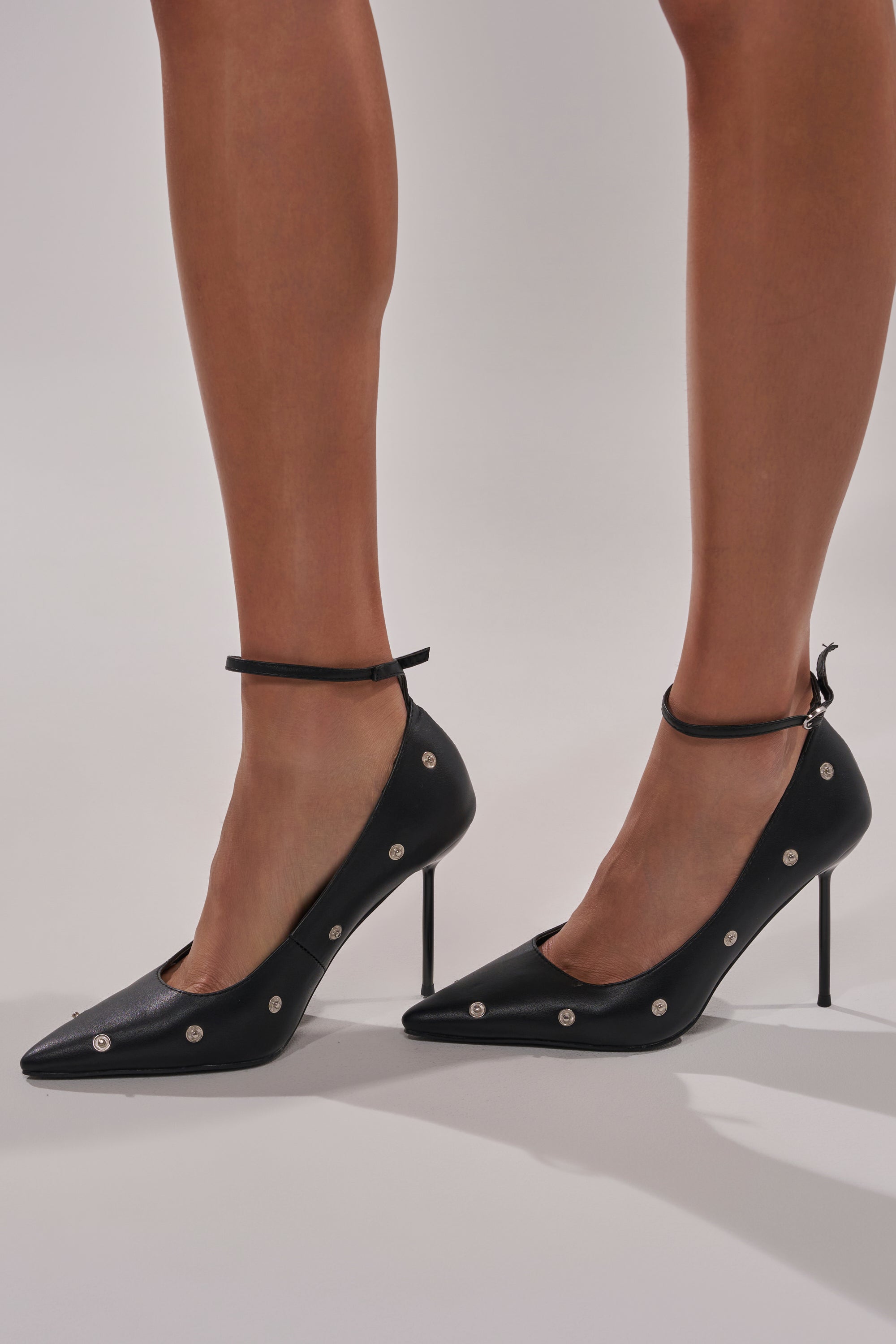 Someone stands on a light-colored surface wearing the AZALEA WANG IMOGALE BLACK SILVER HARDWARE STILETO PUMP featuring pointed toes, ankle straps, and silver stud accents.