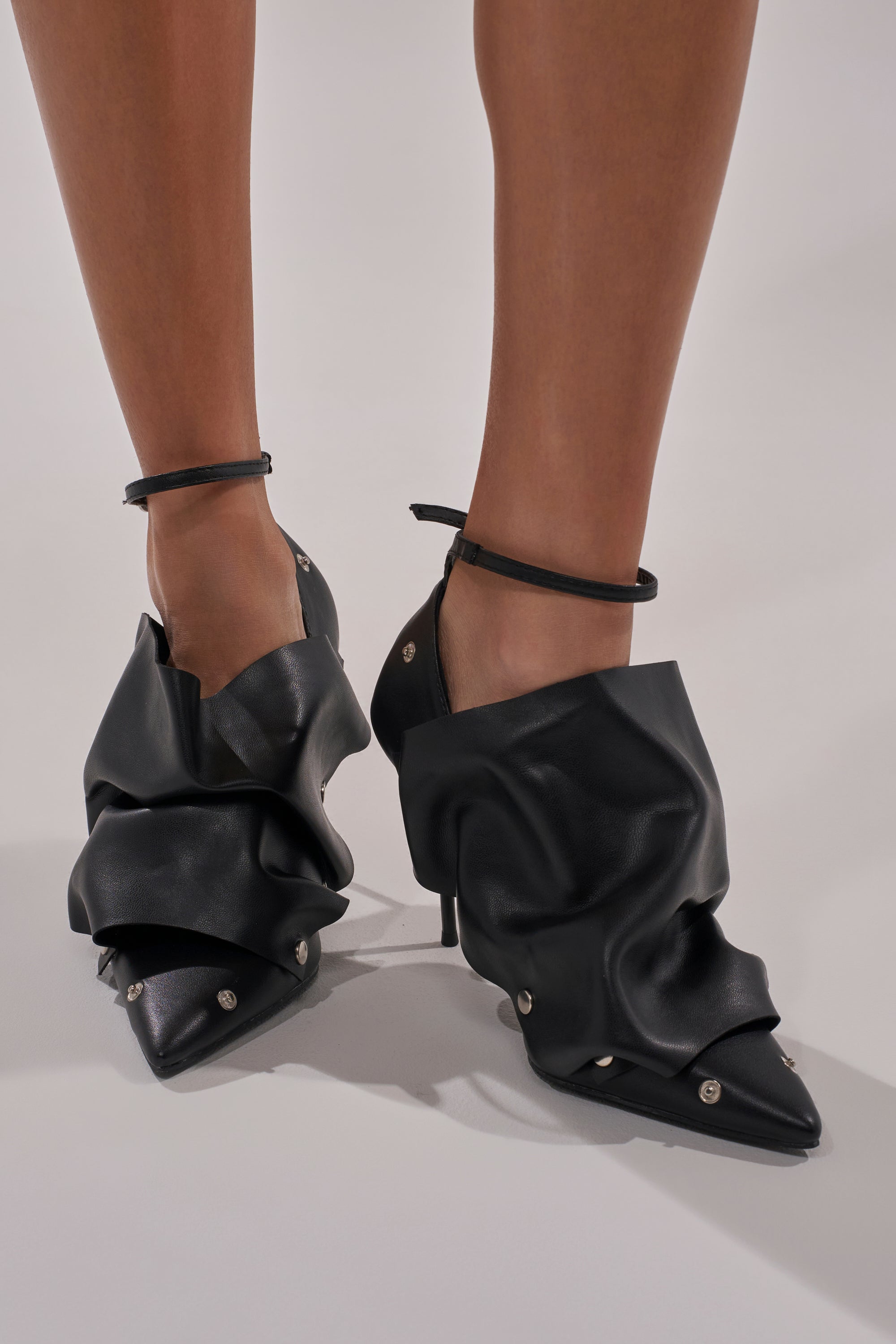 A person wearing AZALEA WANG IMOGALE BLACK SILVER HARDWARE STILETTO PUMPS with draped, ruched leather, metal studs, thin ankle straps, and a pointed toe for an avant-garde look.