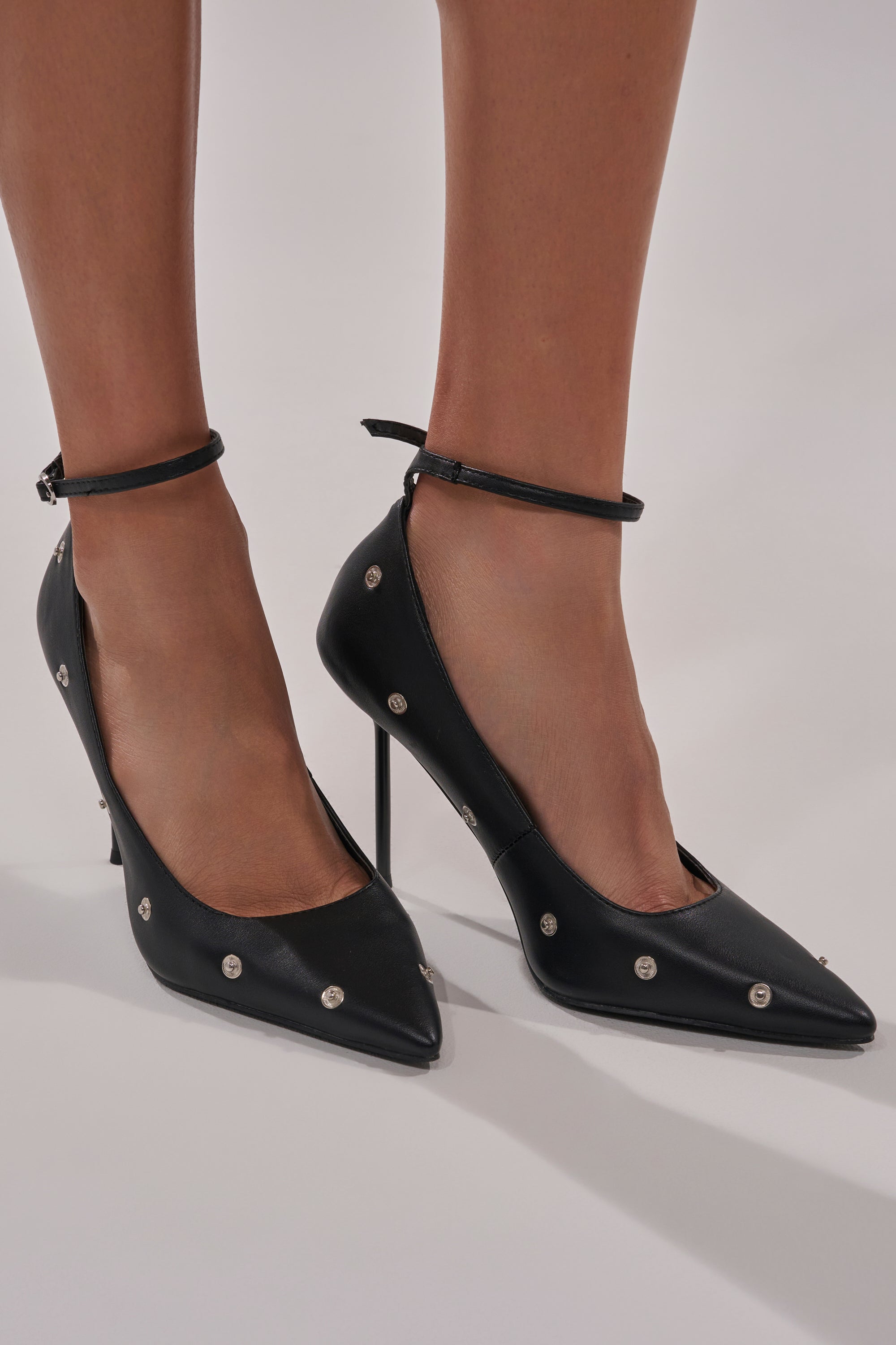 A close-up of a person wearing the AZALEA WANG IMOGALE BLACK SILVER HARDWARE STILETTO PUMP, featuring black pointed toes, ankle straps, and silver studs, as they stand on a light-colored surface.