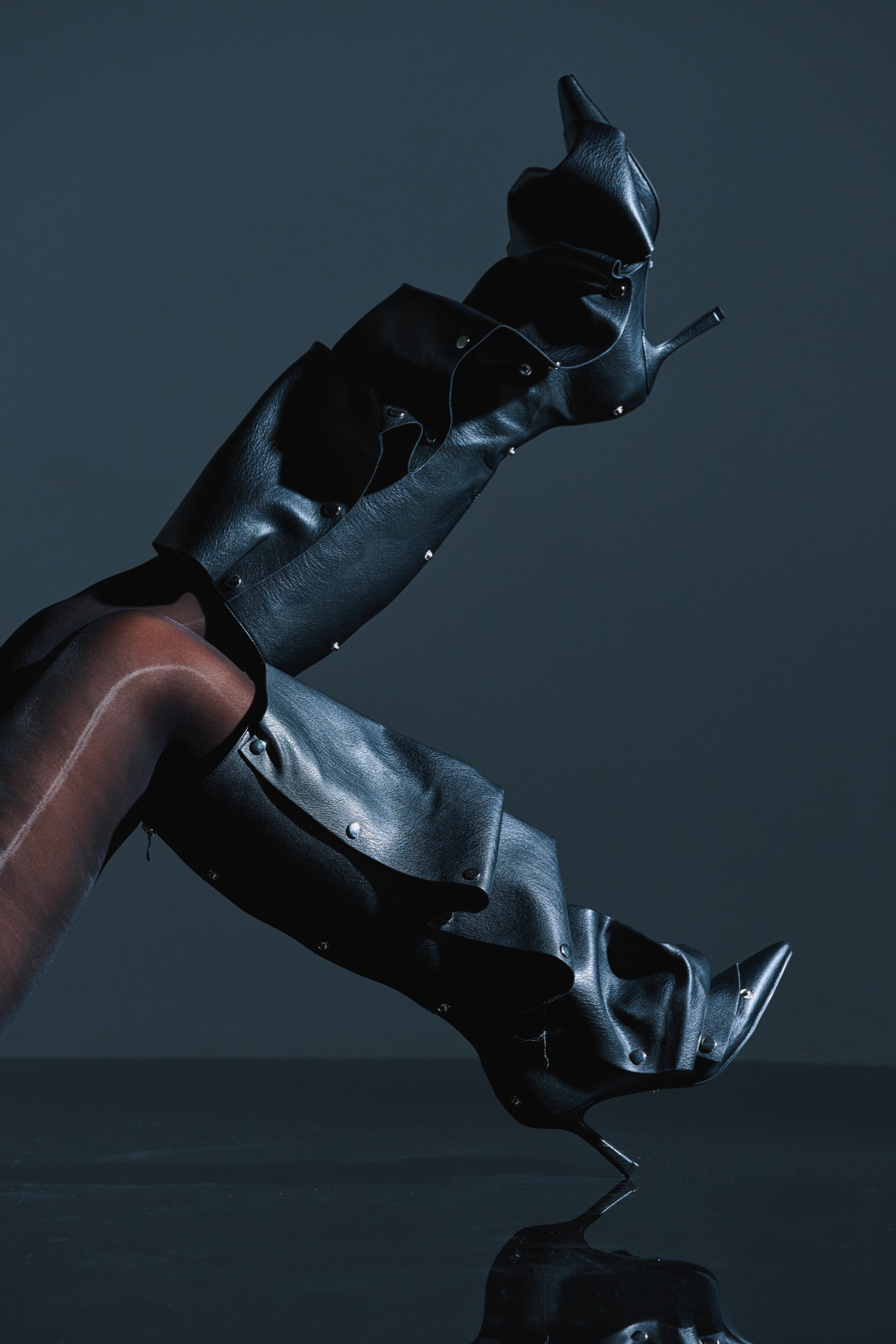 Wearing the AZALEA WANG IMOGINE BLACK SNAP BOOT—sculptural, faux leather knee-high boots with metal studs and stiletto heel—a person poses with one leg raised against a dark background, creating a bold and stylish silhouette.