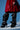 A person wears a shiny red coat and striking AZALEA WANG IMOGINE BLACK SNAP BOOTS—sculptural, knee-high with ruffled layers and a stiletto heel—while standing against a blue background.