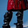 A person wears a shiny red coat and striking AZALEA WANG IMOGINE BLACK SNAP BOOTS—sculptural, knee-high with ruffled layers and a stiletto heel—while standing against a blue background.