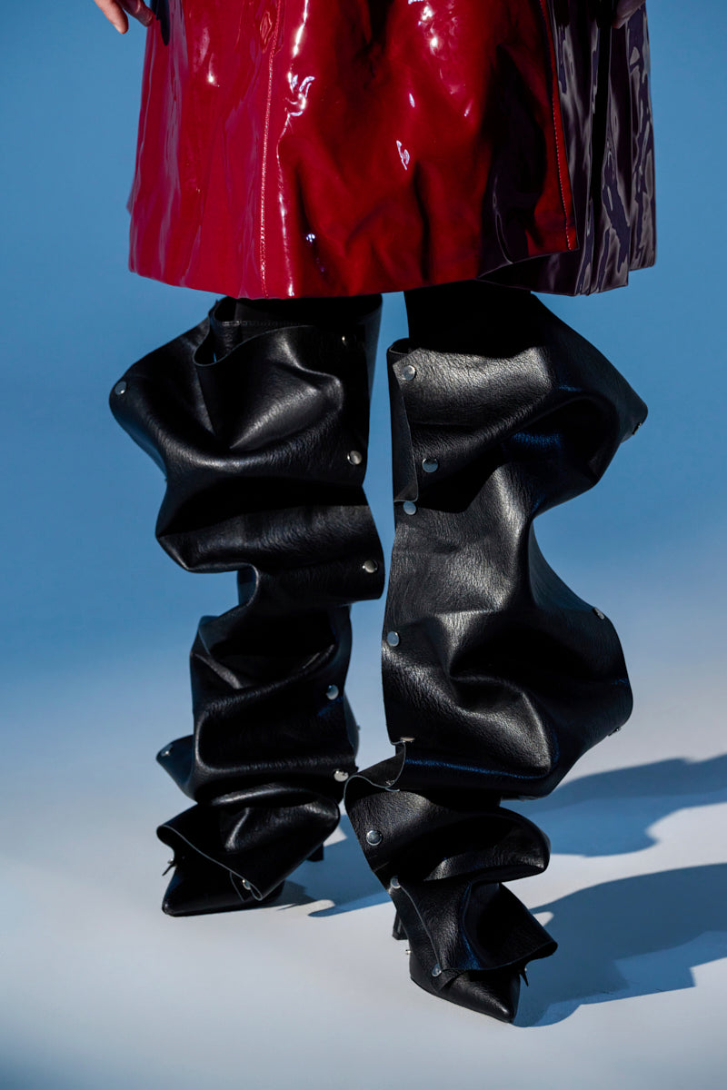 A person in a glossy burgundy coat and black, sculptural, origami-style pants with folds and metal rivets wears AZALEA WANG IMOGINE BLACK SNAP BOOT, standing against a blue background.