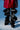 A person in a glossy burgundy coat and black, sculptural, origami-style pants with folds and metal rivets wears AZALEA WANG IMOGINE BLACK SNAP BOOT, standing against a blue background.