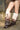 Only the person's legs are visible as they stand on a sidewalk in front of a brick wall, wearing the AZALEA WANG IMOGINE BROWN MULTI FOLDED DETAIL STILETTO BOOT—knee-high, faux leather with ruffles, metallic buttons, and a two-tone gradient.