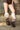 A person wearing high-waisted brown faux leather shorts with large silver studs and AZALEA WANG IMOGINE Brown Multi Folded Detail Stiletto Boots stands on a concrete sidewalk in front of a brick wall.